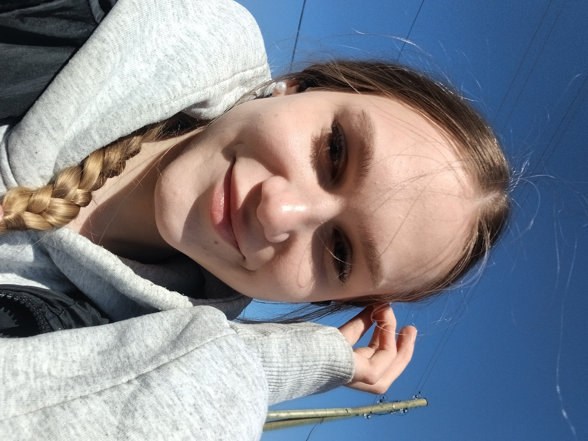 A close-up, tilted portrait captures a young person, likely female, smiling gently. The subject looks upwards towards the camera, her face partially framed by wispy, light brown hair. Her hair is styled in a thick braid that rests over her left shoulder, visible beneath a light gray hooded sweatshirt, worn over a dark garment. She wears a small, white pearl stud earring in her right ear.
The person's expression is pleasant and content. Bright daylight illuminates her face, highlighting her features and casting soft shadows, indicating a sunny daytime setting. She appears to be outdoors under a clear, vibrant blue sky. Faint, thin lines, possibly power lines, are visible stretching across the sky.
In the lower right portion of the image, near the subject's left hand, a light-colored, slender pole or stick is visible, wrapped with small, dark beads or wires. The scene captures a casual, sunny outdoor moment in Bolshaya Sosnova, Russia. There is no visible text in the image.