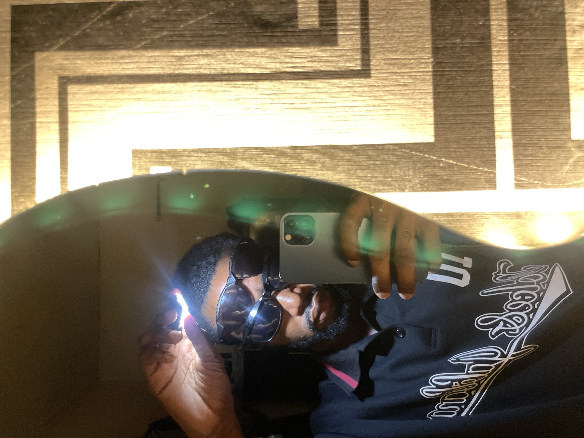 An individual, appearing to be male, with dark skin, a beard, and short dark hair, is depicted in a selfie captured via a mirror reflection within the city of Lokoja, Nigeria. The subject is wearing two pairs of dark sunglasses, one stacked above the other, and a black polo shirt with a pink accent on the collar, featuring white script lettering and the numeral "01" on the chest. The subject holds a grey smartphone, equipped with a triple camera system, in their right hand, pointed towards the mirror. Concurrently, their left hand holds a small, rectangular, bright white light source above their forehead, illuminating their face and hair. The mirror is irregularly shaped on its left edge. To the right of the mirror, a textured surface with a warm, golden hue displays a repeating geometric pattern of interlocking rectangular shapes, possibly indicative of a decorative wall panel or door, exhibiting vertical striations consistent with wood grain. Green lens flares are visible within the mirror's reflection on the left side.