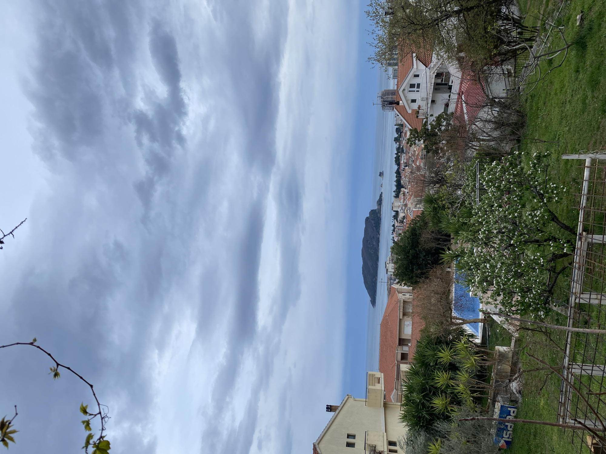 The image captures a view of Budva, Montenegro, taken from an elevated position. The foreground includes residential buildings with red-tiled roofs and lush greenery, including trees and bushes. In the distance, the Adriatic Sea is visible, with the island of Sveti Nikola appearing prominently. The sky is overcast with heavy clouds. A construction site is present with a crane at work. The image offers a glimpse into the coastal landscape of Budva, blending urban elements with natural features.