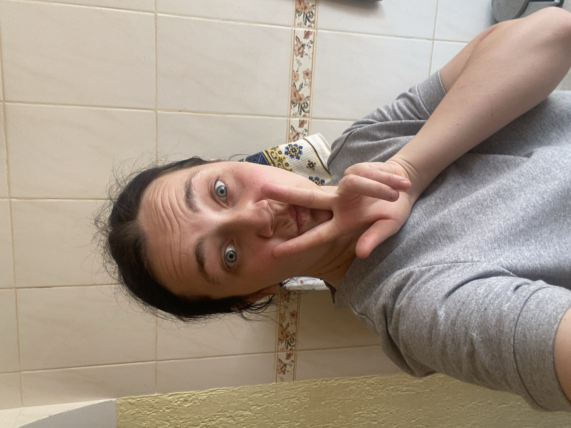 A close-up selfie shows a young woman with dark hair, bright blue eyes, and fair skin. She is wearing a gray t-shirt and is making a "peace" sign with her right hand in front of her face. Her mouth is slightly open, as if she is about to speak or pucker her lips. The background appears to be a tiled wall with a decorative border. A towel with a blue and yellow pattern hangs on the wall behind her left shoulder. The lighting is bright and appears to be natural. The woman is positioned in the center of the frame.