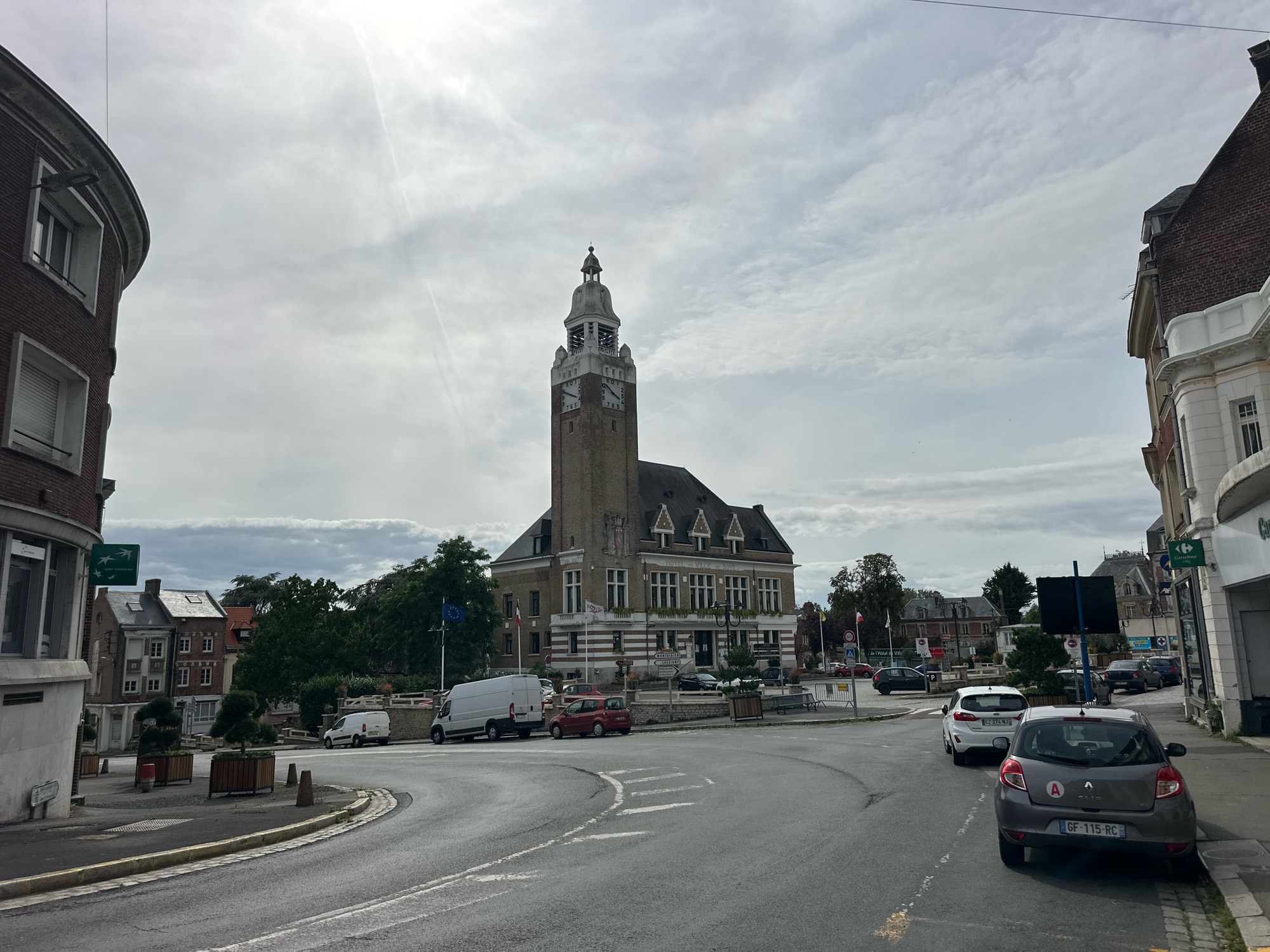This image shows a street scene in the city of Roye, France, featuring a large, brick building with a prominent clock tower. The building appears to be a public structure, possibly a town hall, with the words "HOTEL DE VILLE" visible on its facade. Several cars are parked on the street, including a white van and a grey Renault Clio. In the background, there are other buildings, trees, and a cloudy sky. A street sign with the text "ROYE" is visible, confirming the location. The overall impression is of a typical European town square on an overcast day.