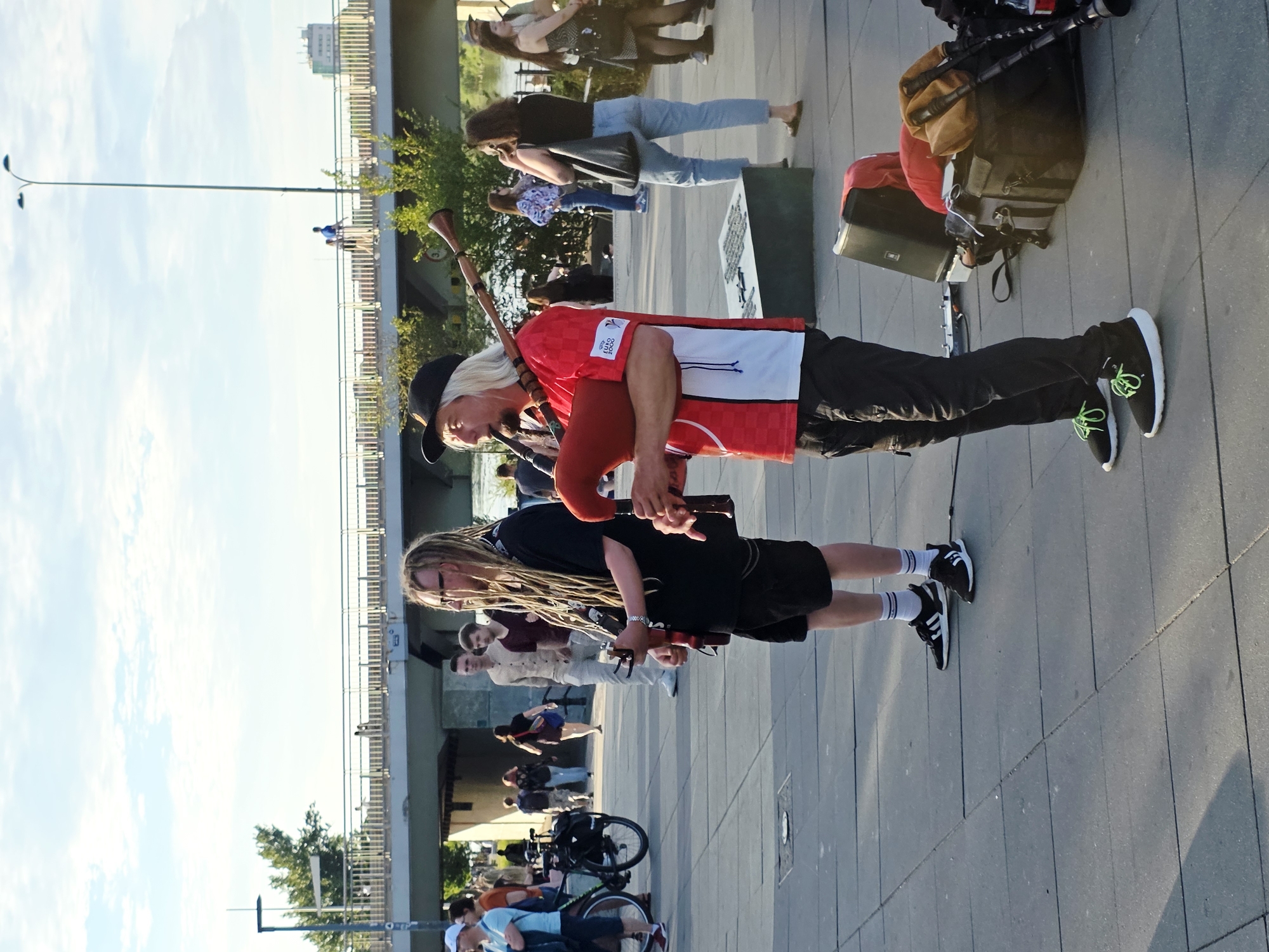 Two male musicians are performing bagpipes on a paved plaza in Warsaw, Poland, with a bridge and a body of water in the background. The musician on the left, with long dreadlocks, wears a black t-shirt, black shorts, white socks with blue stripes, and black sneakers. He is holding a set of bagpipes. The musician on the right, wearing a black baseball cap and a red and white shirt, is playing his bagpipes. He has blonde hair and is wearing black pants and black sneakers with green laces. Various bags and equipment are visible on the ground near the musicians. In the background, people are walking on the plaza and a bridge spans over the water. Trees and a lamppost are also visible in the scene. The sky is partly cloudy.