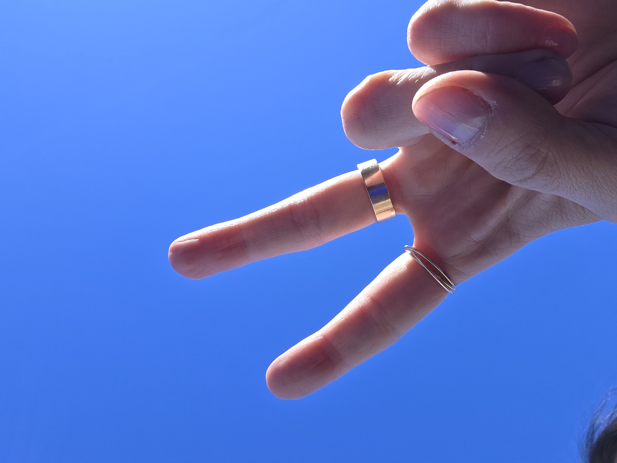 The image depicts a hand making a peace sign against a clear blue sky. The index finger has a thick gold band, and the pinky finger has two thin silver rings. The person's hair is visible at the bottom right corner.