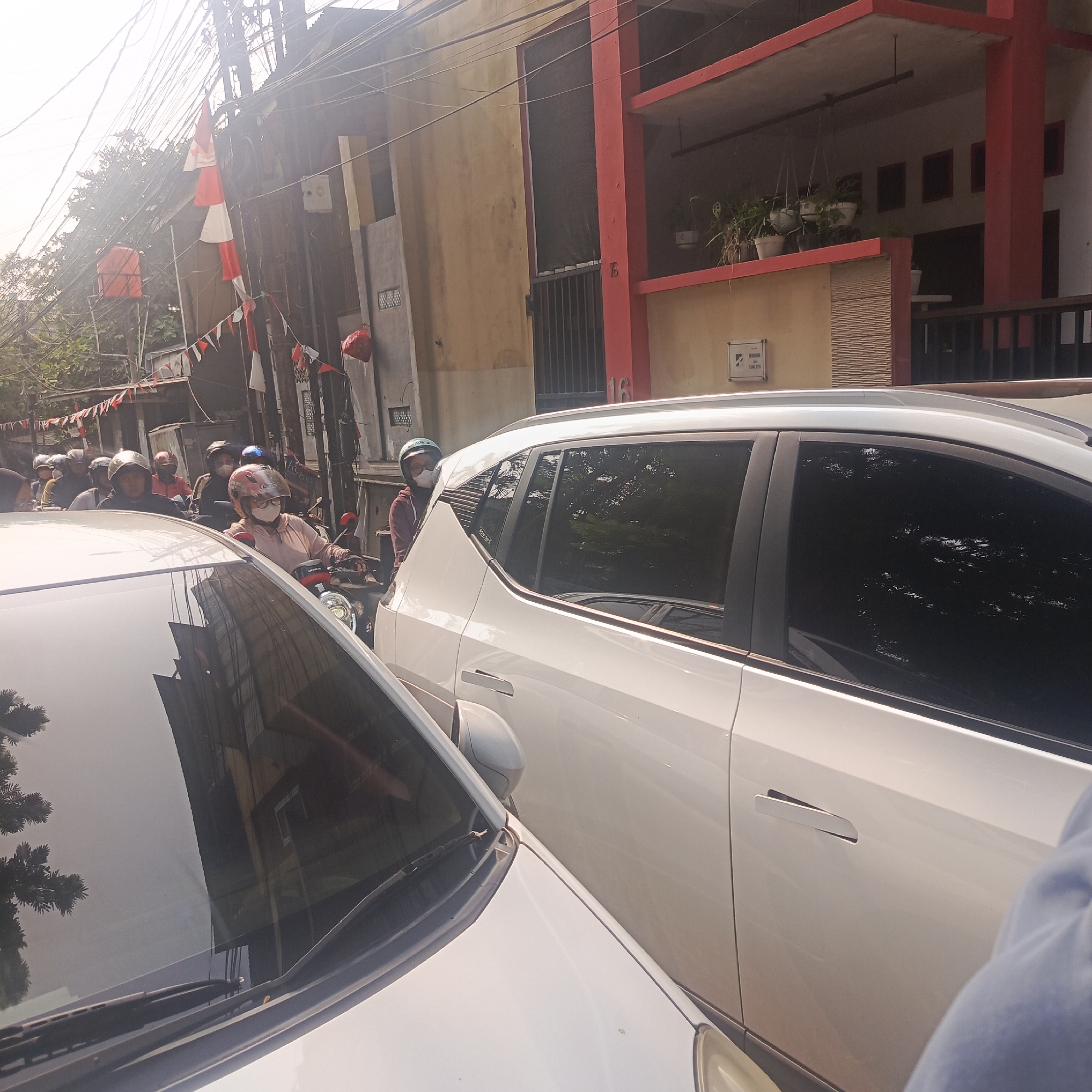 This is a photograph taken on a street in South Jakarta, Indonesia, during daylight hours. The scene depicts a traffic jam with numerous motorcycles and at least two cars. On the left side of the image, a white car's windshield and hood are prominently featured, with reflections of trees and buildings visible on the windshield. To the right of this car, a white SUV is parked, showing its side profile with tinted windows and a visible door handle. A person wearing a blue jacket is partially visible on the right edge of the frame, seemingly exiting or interacting with the white SUV.
In the background, a dense collection of motorcycles is evident, with many riders wearing helmets and some wearing face masks, suggesting the presence of COVID-19 precautions. The architecture of the buildings lining the street includes a two-story structure with red accents and balconies, as well as a yellow building. Electrical wires are strung across the scene, typical of urban Indonesian environments. Decorative red and white flags, possibly representing the Indonesian national colors, are strung along poles and wires, suggesting a local celebration or event. The overall atmosphere suggests a busy, congested urban street environment.