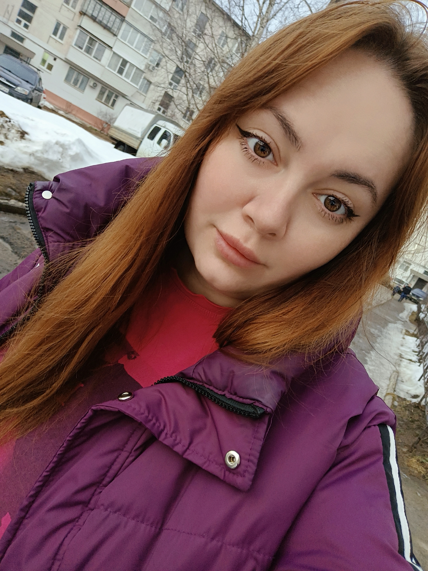 The image depicts a young woman taking a selfie outdoors in what appears to be a residential area of Chaikovsky, Russia, during late winter or early spring. The woman, positioned prominently in the foreground, has long, straight, reddish-brown hair, brown eyes with winged black eyeliner, and a neutral expression. She is wearing a dark purple quilted jacket with a black zipper and silver snap buttons, over a red or magenta patterned top. A black stripe with two white stripes is visible on her right jacket sleeve.
The background reveals a street scene characterized by melting snow and wet pavement. On the left, large piles of snow accumulate next to a dark-colored parked sedan and a white commercial van with a utility canopy. Beyond these, a multi-story, light-colored residential building with numerous windows and balconies is visible. Further to the right, patches of bare ground and wet asphalt lead towards other indistinct multi-story buildings and sparse, bare deciduous trees. Two figures are visible walking on the wet pavement in the mid-ground on the right side. The sky is overcast.