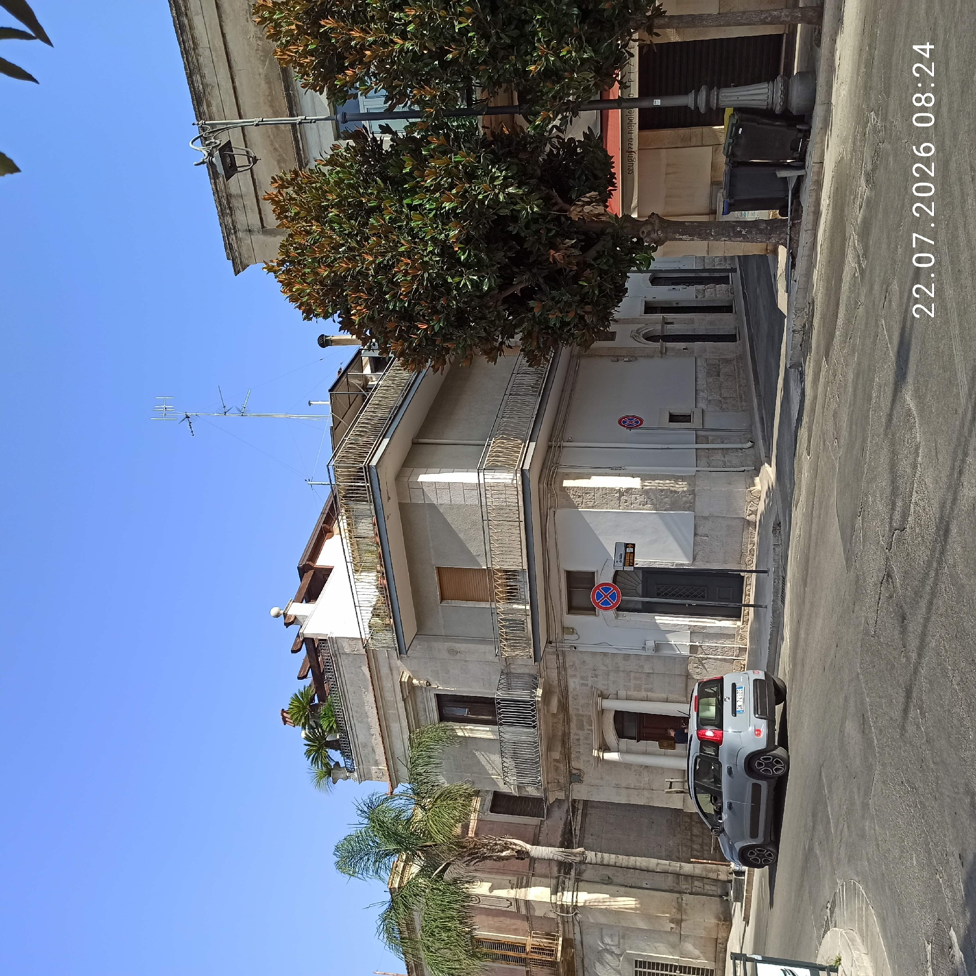 A light gray Fiat Panda is parked on the right side of a street in Modugno, Italy, with its headlights on. The car is facing away from the camera. The street is paved with asphalt and shows signs of wear and tear. On the left side of the street, there is a building with a palm tree in front of it. The building has multiple floors with balconies. On the right side of the street, there are more buildings and trees. In the background, there is a clear blue sky. A date and time stamp, "22.07.2026 08:24", is visible in the bottom right corner of the image.