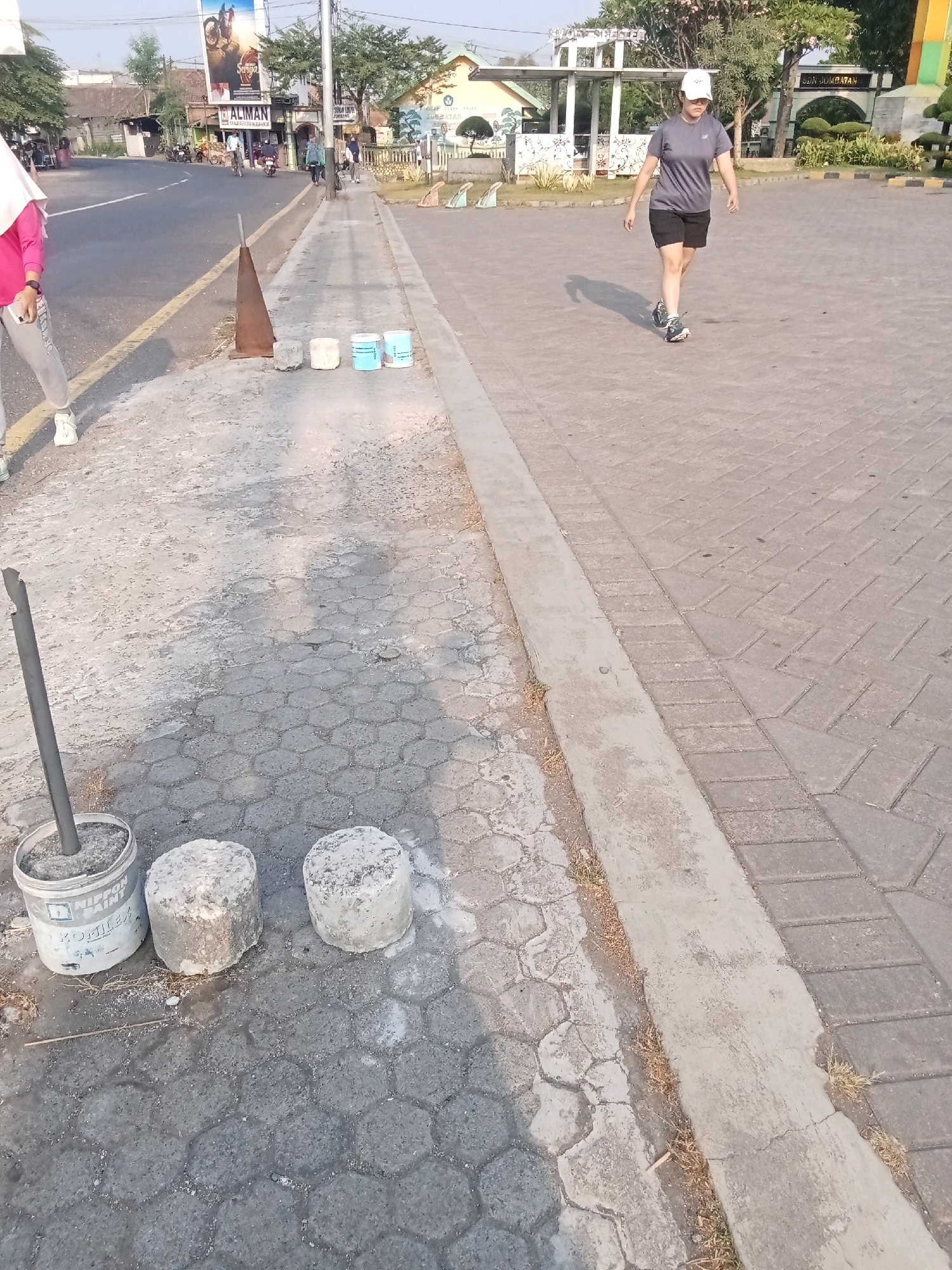 The image depicts a sunny outdoor scene on a paved area in Kaliwungu, Indonesia. In the foreground, on the left side, a person wearing a pink hijab and grey sweatpants is partially visible, with one hand holding a smartphone. Adjacent to this person, a metal pole stands upright in a white bucket labeled "NIPPON PAINT KONTILLEX". Two cylindrical concrete blocks are placed on the hexagonal-patterned pavement next to the bucket.
Moving towards the center and right of the image, a woman in a grey t-shirt, black shorts, and a white baseball cap is walking away from the camera. She is wearing athletic shoes and her shadow is cast on the pavement.
The background shows a street with vehicles, including motorcycles and cars, and several buildings. One building has a prominent billboard with text that appears to be Indonesian. Another building has a sign that reads "SDN JUMBATAN". Trees and other vegetation are visible, along with streetlights. The overall environment suggests a public area possibly near a school or community center.