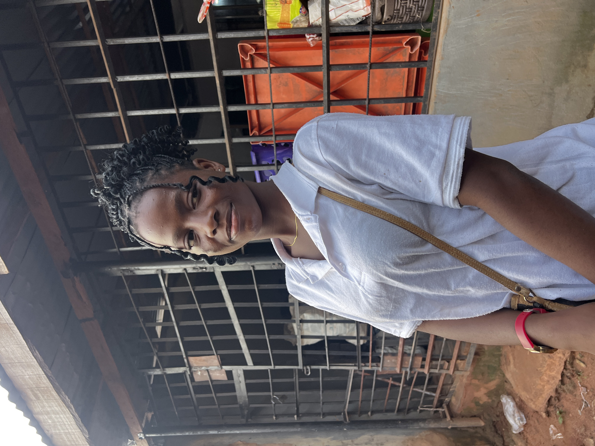 A young Black woman with braided hair and a white collared shirt smiles at the camera. She wears a thin gold necklace and a pink fitness tracker on her left wrist. A tan strap for a bag is visible across her shoulder.
The background appears to be outdoors, possibly in a market or residential area in Nkpor, Nigeria. Behind her, a metal grate structure is prominent, with orange plastic crates and some bags of goods visible through it. The overall lighting suggests it is daytime. The scene is slightly tilted, making the orientation of the image unusual.