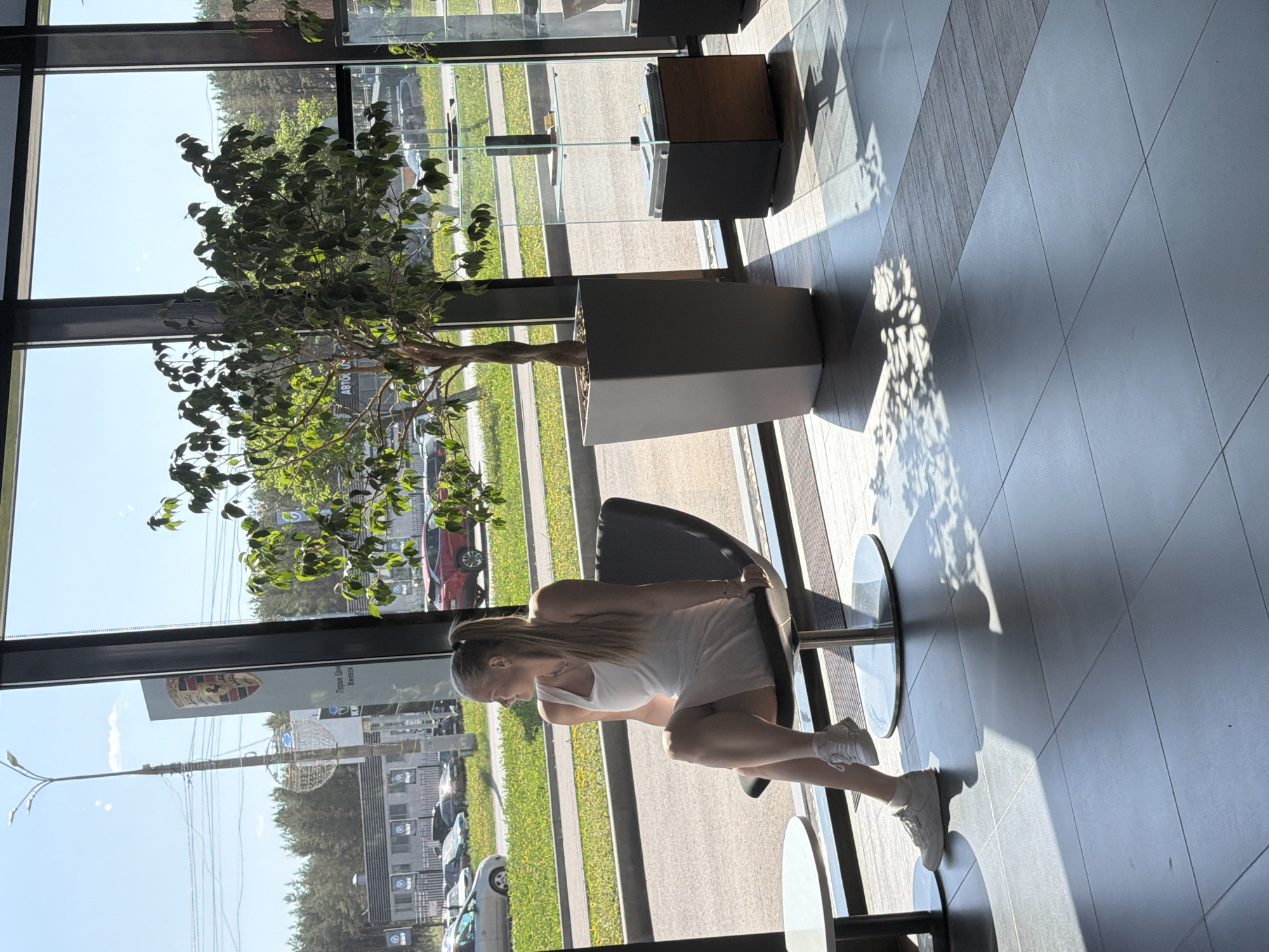 A young woman is seated in a chair inside a building. She is wearing a white tank top and shorts and white sneakers. She is seated near a window. Outside the window, there is a street with cars and buildings. A Porsche dealership can be seen. The building's address is 176B Ulitsa Karla Marksa in Izhevsk, Russia. A tree is in a pot inside the building, near the window. The floor is tiled.