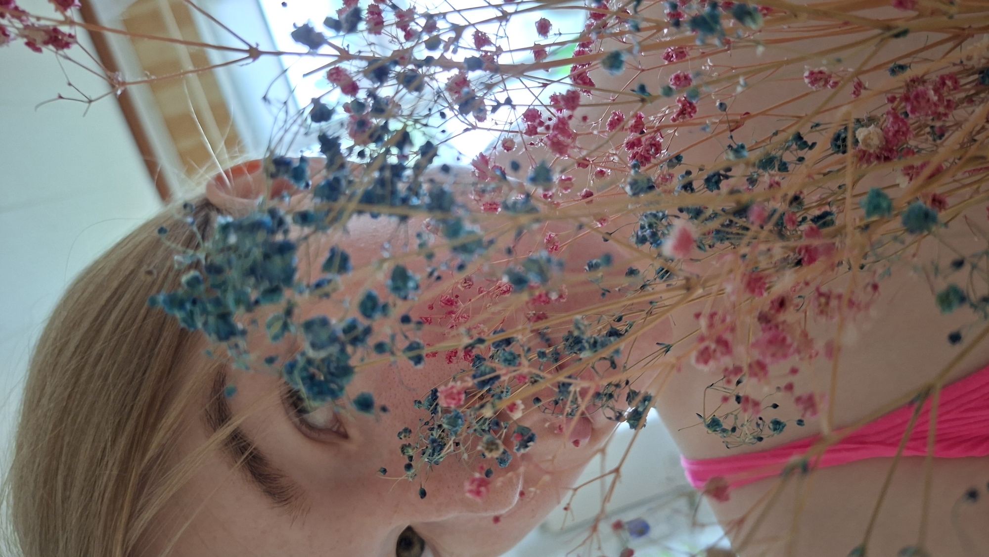 A close-up, low-angle shot focuses on a person partially obscured by dried baby's breath flowers, possibly indoors. The individual has light blonde hair and fair skin, with the visible eye being brown. A pink strap, likely part of clothing, is visible in the lower right. The background includes a window and a light-colored wall. The location, Большой Букор, Russia, is not verifiable from the image content alone.