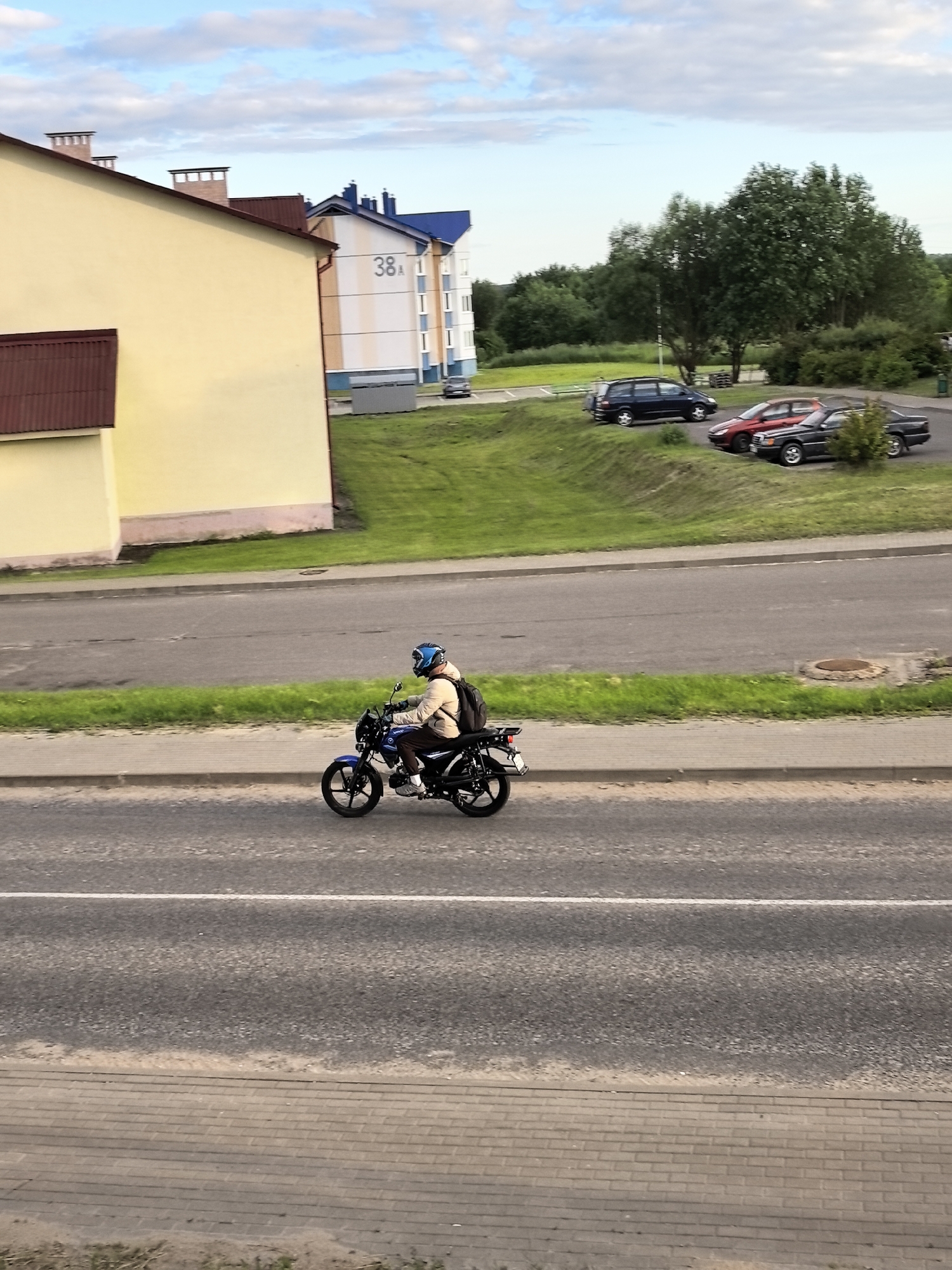 A person rides a blue motorcycle down a paved road in an urban environment. The rider is wearing a tan jacket, dark pants, and a blue helmet. They have a black backpack on their back. The motorcycle is a scooter-style with black wheels and accents. The road has a white line painted down the center. To the left of the road is a sidewalk made of brick pavers, and beyond that is a grassy slope and a yellow building. To the right of the road is a grassy area with trees and parked cars, including a dark blue minivan, a red sedan, and a black sedan. In the background, there are apartment buildings with blue roofs and a sign that reads "38A". The sky is blue with scattered clouds.