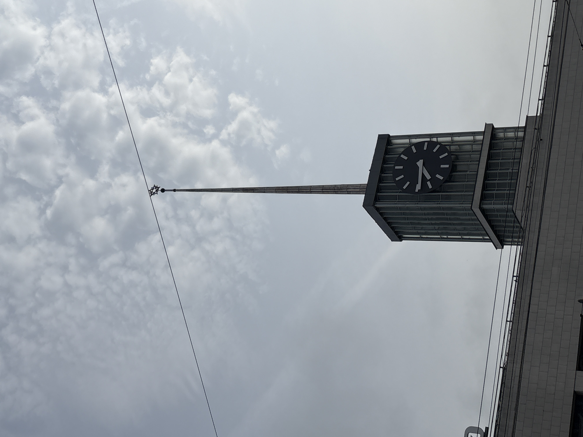 The image shows a clock tower with a long spire, likely located in Saint Petersburg, Russia. The sky is overcast with light gray clouds. A thin cable runs diagonally across the frame, intersecting with the spire near its top where a star-shaped finial is located. The clock face is dark with light-colored numerals and hands, showing the time as roughly 7:05. The tower itself is multi-tiered, with a grid-like structure visible below the clock face. A portion of the adjacent building with light gray tiles is also visible.