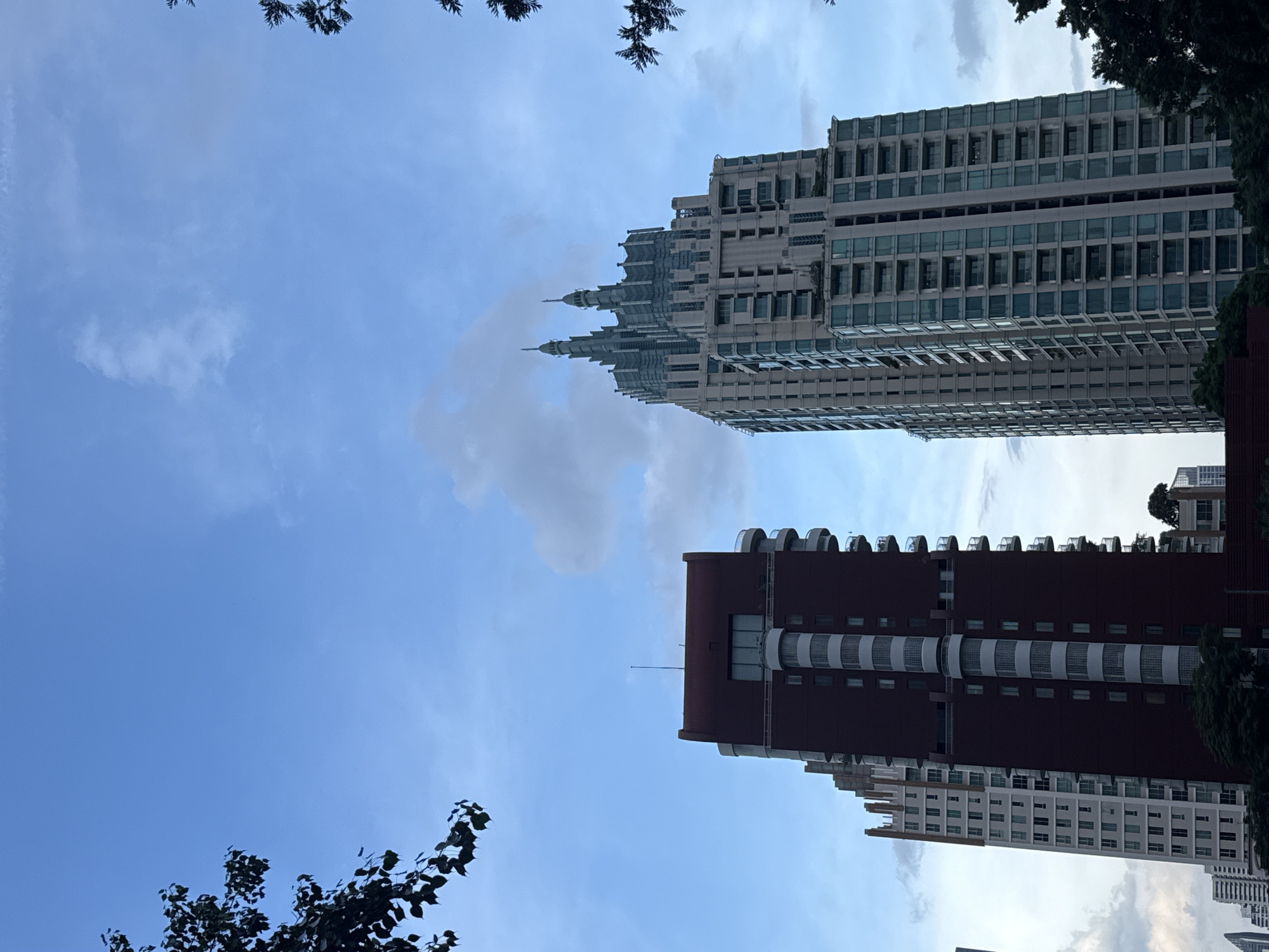 The image captures a low-angle view of modern skyscrapers in Bangkok, Thailand, set against a sky with scattered clouds. On the left, a dark red, rectangular building with white circular balconies dominates the foreground. To its right, a taller, light-colored building with a more ornate, stepped design featuring two spires rises dramatically. The facades of both buildings are primarily glass and concrete, indicative of contemporary urban architecture. Trees with dark green foliage are visible framing the top and bottom edges of the frame, suggesting a tree-lined street or park area in front of the buildings. The scene appears to be captured during daylight, likely in the late afternoon or early evening, given the soft lighting and the presence of clouds.