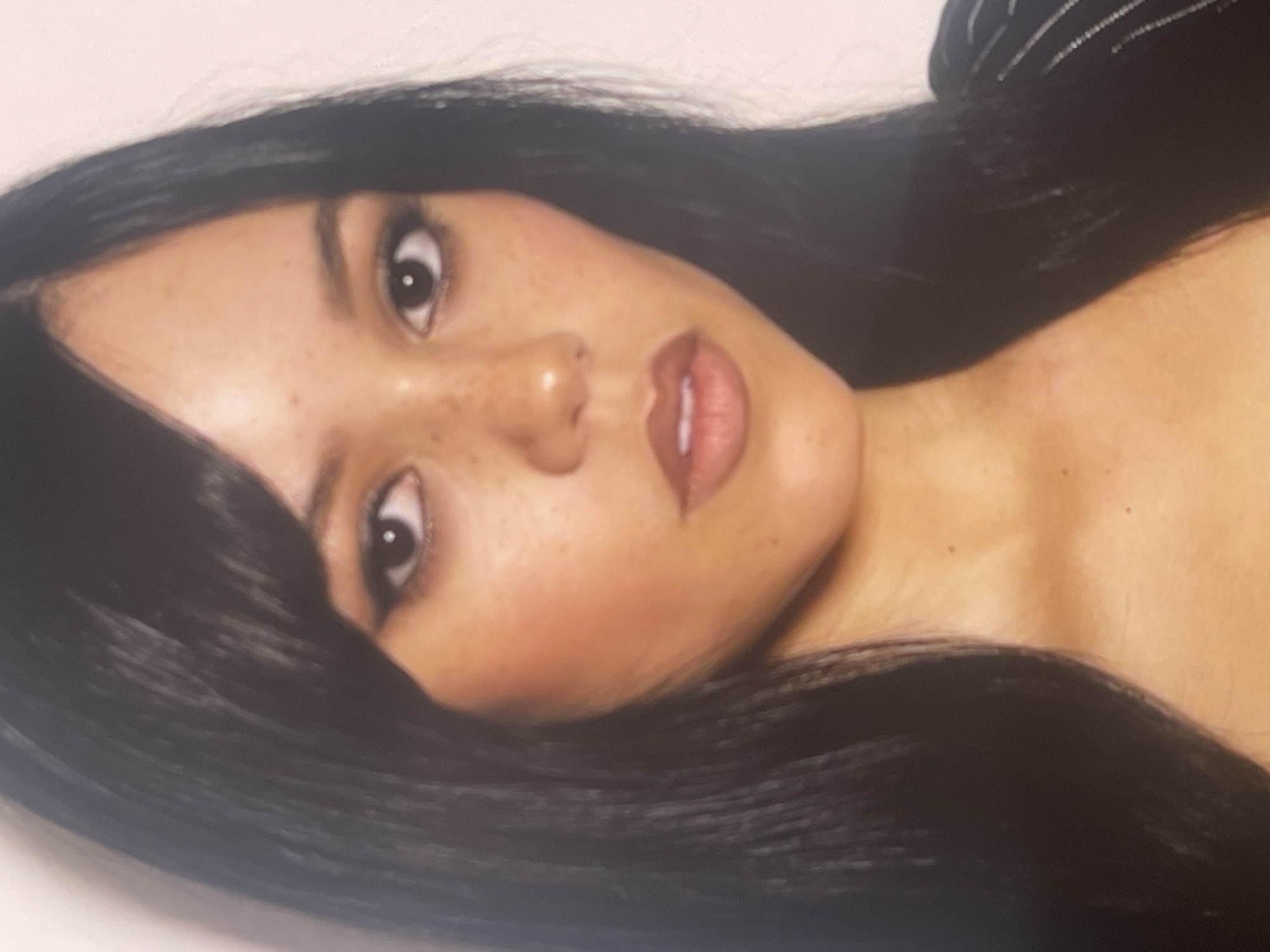 The image features a close-up portrait of a young woman, presented with her head positioned towards the left side of the frame due to the image's orientation. She has long, straight, dark hair that frames her face and extends upwards. Her light-toned skin is adorned with numerous small freckles visible across her nose and cheeks. Her dark eyes, accentuated by prominent dark eyeliner, gaze directly towards the viewer. She wears blush, giving a rosy tint to her cheekbones, and her lips are painted with a reddish-brown, slightly glossy lipstick, parted just enough to show a hint of her upper teeth. A small part of a dark garment with subtle light stripes is visible in the upper right portion of the frame, hinting at a top or dress. The background is a plain, light color, offering no specific environmental details. There is no visible text. The woman's expression is direct and composed. This photo is set in Poltava, Ukraine.
