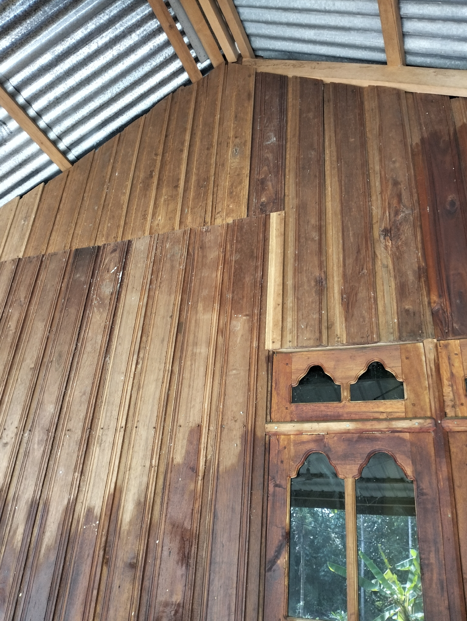 The image shows a portion of a wooden structure, possibly a house or a shed, with a corrugated metal roof. The walls are constructed from vertical wooden planks of varying shades of brown, with some exhibiting a wet or varnished appearance. There are two windows visible, set within wooden frames. The upper window has an unusual decorative top with two arch-like shapes. The lower window is divided into two panes and reflects green foliage, suggesting the presence of trees or plants outside. The roof is supported by wooden beams. The overall scene depicts an outdoor, rustic environment in Mandala Jaya, Indonesia. No people are present.