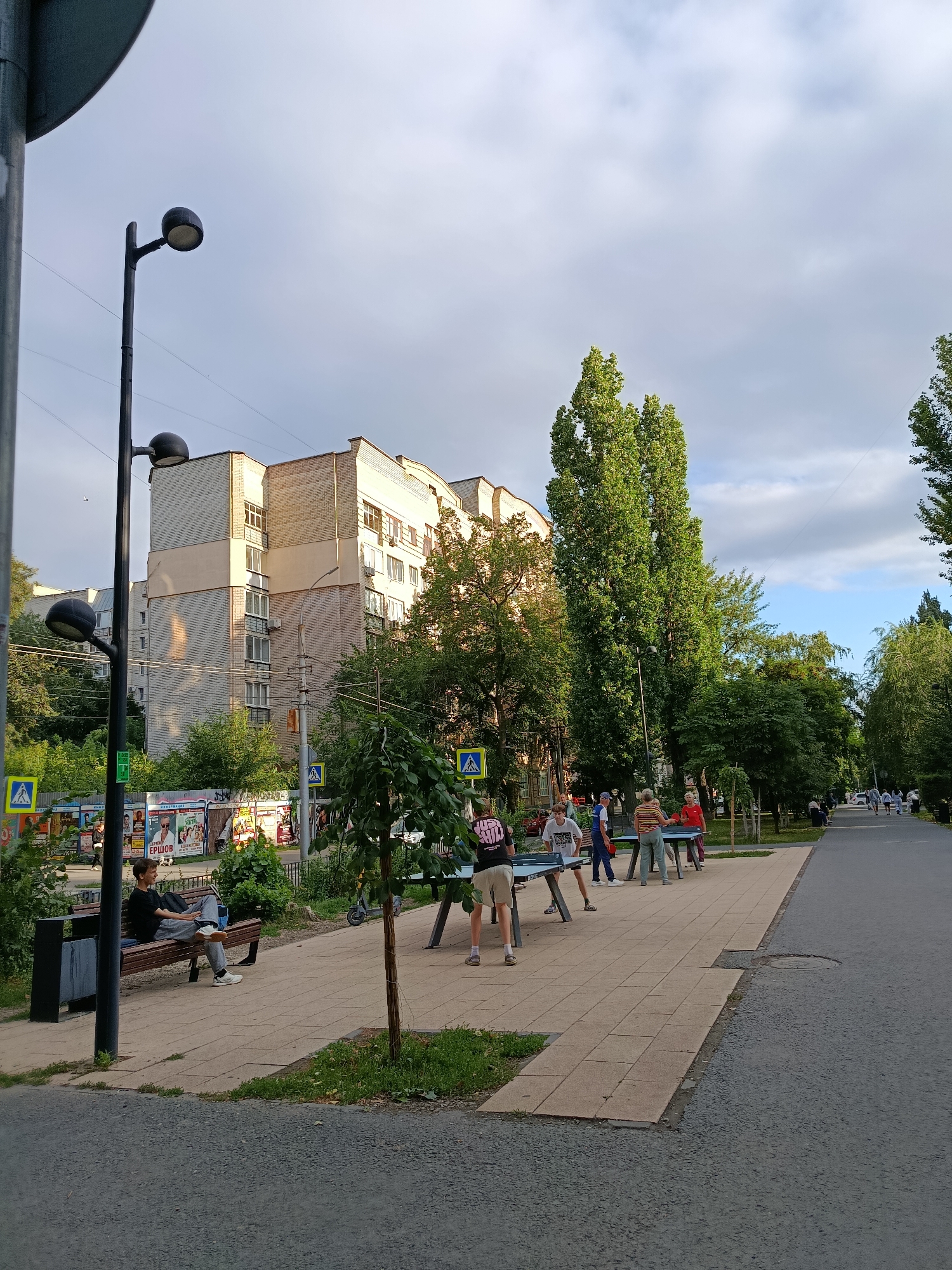 A street scene in Saratov, Russia, captures people engaged in various activities on a warm day. In the foreground, a person is seated on a park bench, appearing relaxed. Further down the paved walkway, a group of individuals are actively playing table tennis, with several tables set up. The background reveals a multi-story brick apartment building and lush green trees lining the street. Traffic signs and advertisements are visible, indicating a public urban area. The sky is partly cloudy, with hints of blue sky.