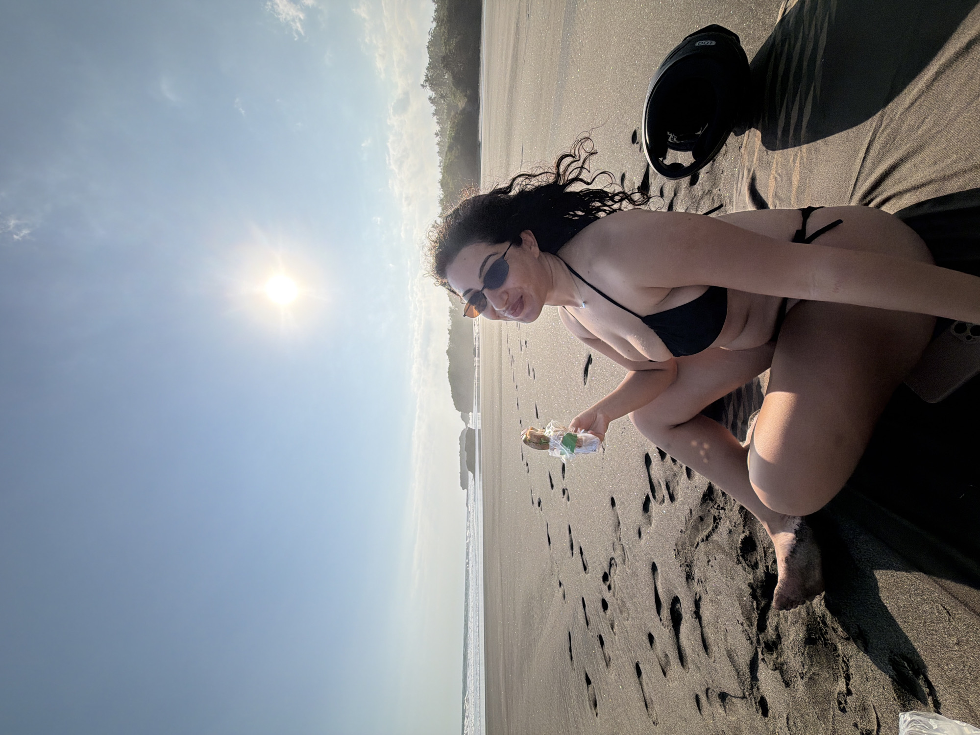 The image displays an adult female seated on a dark sand beach in Selemadeg, Indonesia. She has dark, curly hair and wears a black string bikini and dark, slim sunglasses. She is seated cross-legged on a dark fabric, looking towards the camera with a slight smile while holding a clear plastic-wrapped item, possibly a snack or beverage, which features a green element on its packaging. Her bare feet are visible, partially covered in sand. To her right, a black helmet rests on the dark sand, casting a distinct shadow. The background reveals a wide expanse of dark sand with numerous footprints, leading to the ocean where white-capped waves are breaking. Distant land formations or cliffs, covered in green tropical vegetation, are visible beyond the water. The sky is bright blue with minimal scattered clouds, and the sun is prominently visible in the upper left, producing strong glare and casting long shadows across the beach.
