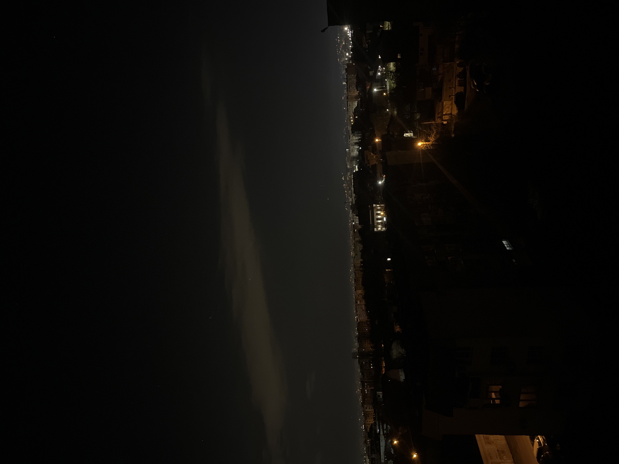 The image shows a night view from a high vantage point in a city. The left side of the frame is dominated by a dark expanse, likely a body of water. The horizon is visible in the distance. The right side of the frame reveals a cityscape with numerous buildings and lights, indicating urban development. Streetlights are interspersed amongst the buildings. Due to the darkness and angle of the shot, it's difficult to discern more specific details about the structures and environment, but the lights and urban configuration suggest a populated area. The absence of signs, distinctive landmarks, or other clear identifiers makes it difficult to identify the location as Sofia, Bulgaria. There are no people in the frame. The interaction between the city lights and darkness is notable, with the lights providing the primary source of illumination in the urban area.