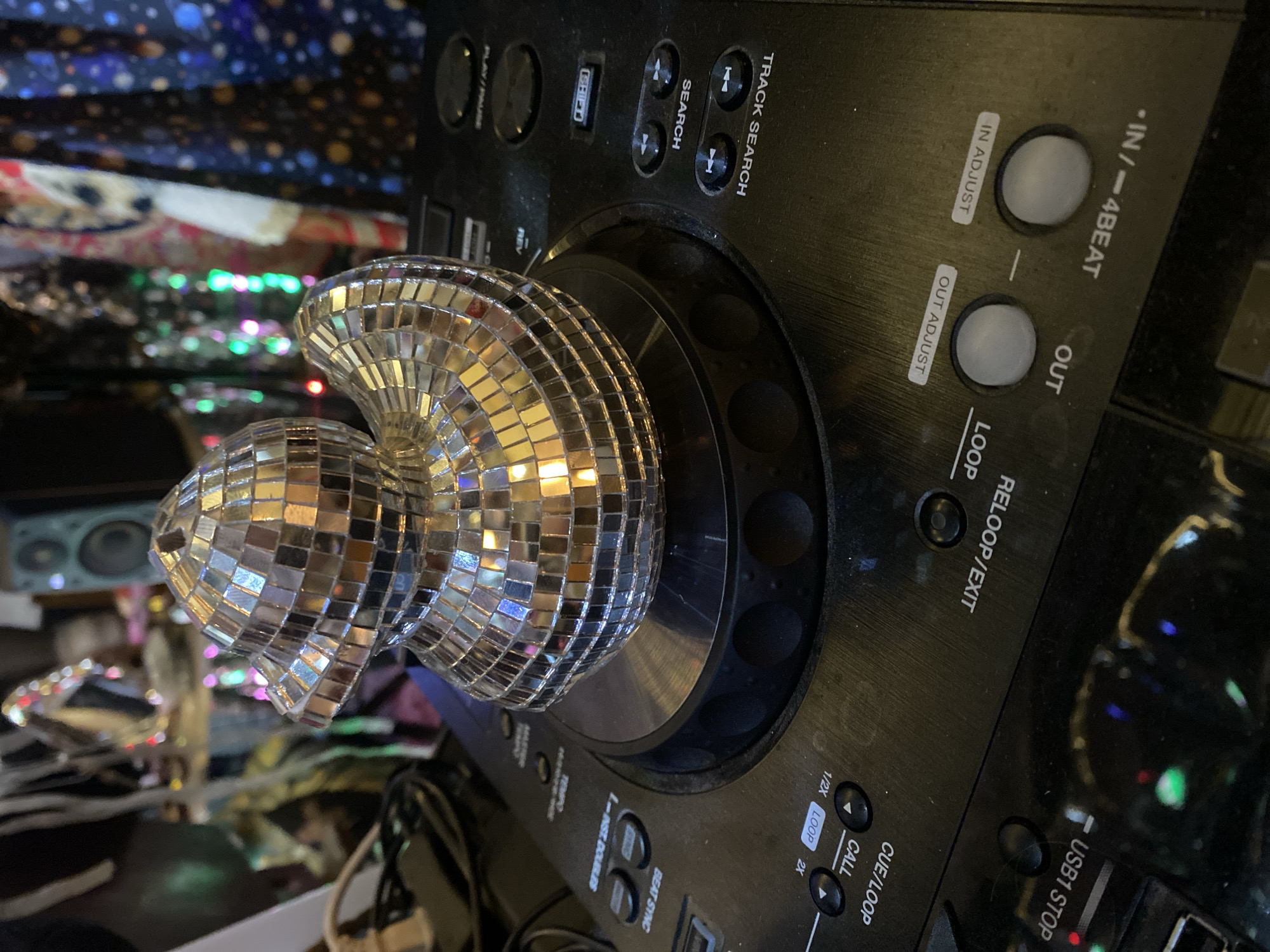The image shows a DJ controller with a heart-shaped disco ball resting on the turntable. The controller is black and has various buttons and knobs. Text visible on the controller includes "TRACK SEARCH", "LOOP", "USB1 STOP", "CUE/LOOP", "CALL", "BEAT SYNC", and "TEMPO". The disco ball is made of small mirrored squares, reflecting light. The background is cluttered with various objects including speaker, LED lights, cloth and cables. The image does not contain information to verify it was taken in Baan Nai Suan, Thailand.