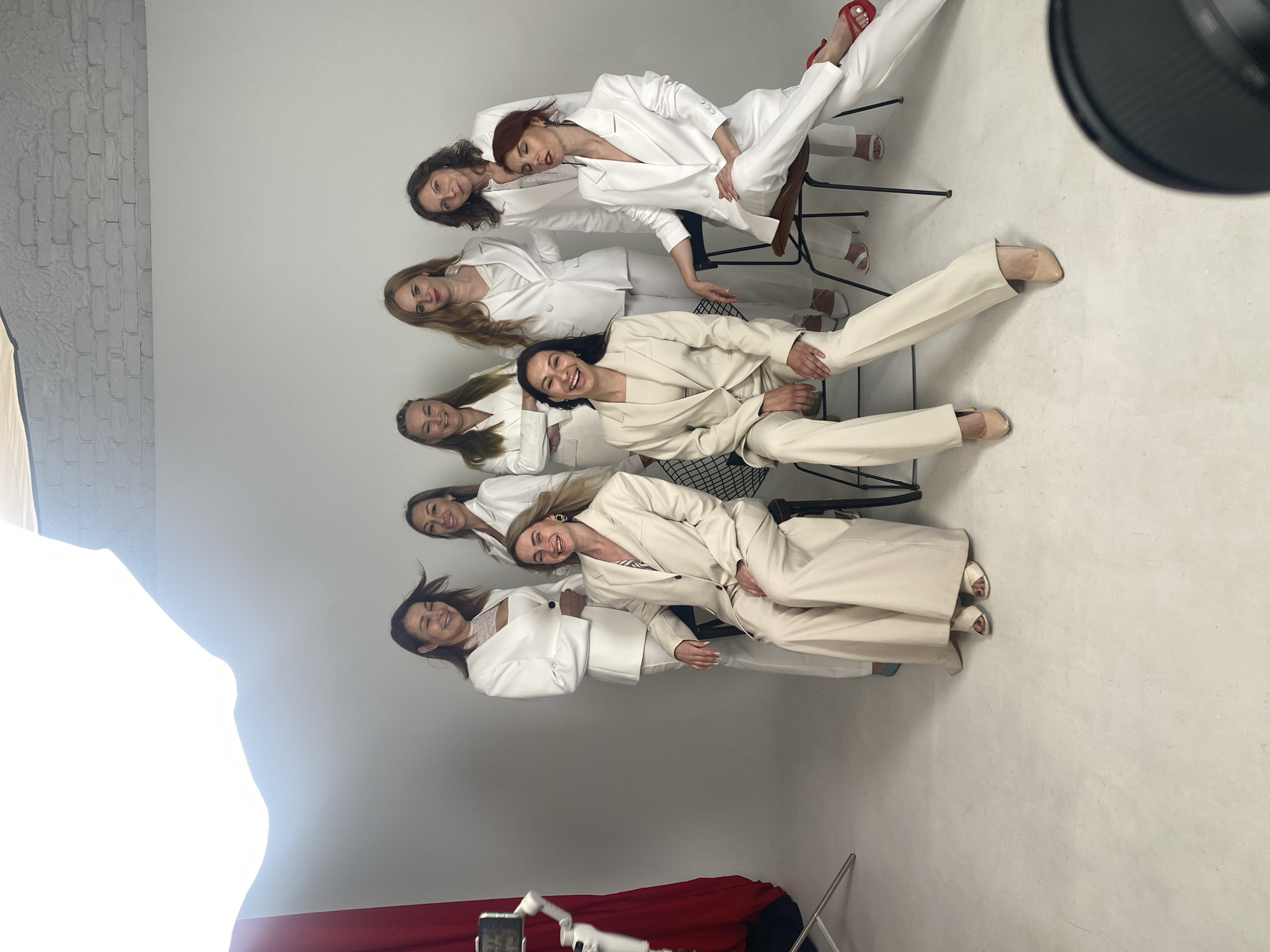 Eight women are posing in white or off-white pant suits against a white studio backdrop. The lighting is bright and diffused. A red curtain is visible on the left side of the image. A camera on a gimbal is also present on the left. Some of the women are standing and some are seated on chairs. They are all looking towards the camera and appear to be smiling or laughing. The setting is a professional photography studio, likely in Sochi, Russia, given the request.