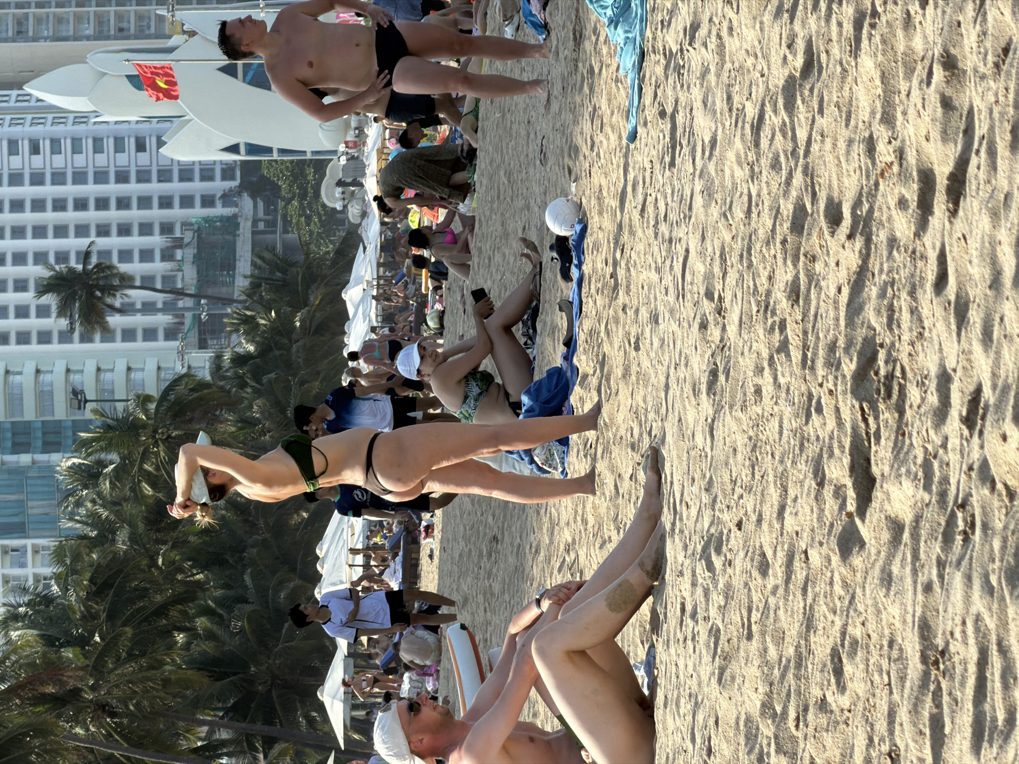 The image captures a lively beach scene in Nha Trang, Vietnam, under bright, clear skies, likely during late morning or early afternoon as evidenced by the distinct shadows on the sand.
In the foreground, on the left, a shirtless man in black swim trunks stands with his hands on his hips, looking towards the right. Behind him and slightly to the left, a large white structure, possibly part of a boat or resort architecture, displays a red Vietnamese flag with a prominent yellow star. Further into the frame, a woman in a dark green bikini top and light-colored bottoms is seen from behind, standing with her arms raised.
Towards the right, a person wearing a white cap and sunglasses reclines on the sand, propped up on a striped beach cushion, with legs extended. Nearby, another individual in a white cap sits on the sand, holding a smartphone. A white volleyball and some blue fabric (possibly a towel) lie on the sand.
The midground and background are densely populated with numerous beachgoers. Many are relaxing on the sand, some under white beach umbrellas that dot the shore. Behind the beach, a vibrant line of lush palm trees provides a natural border before a backdrop of towering high-rise buildings, characteristic of an urban coastal setting. The overall atmosphere is one of leisure and vacation on a popular, sun-drenched beach.
Visible text is limited to the yellow star on the red Vietnamese flag.