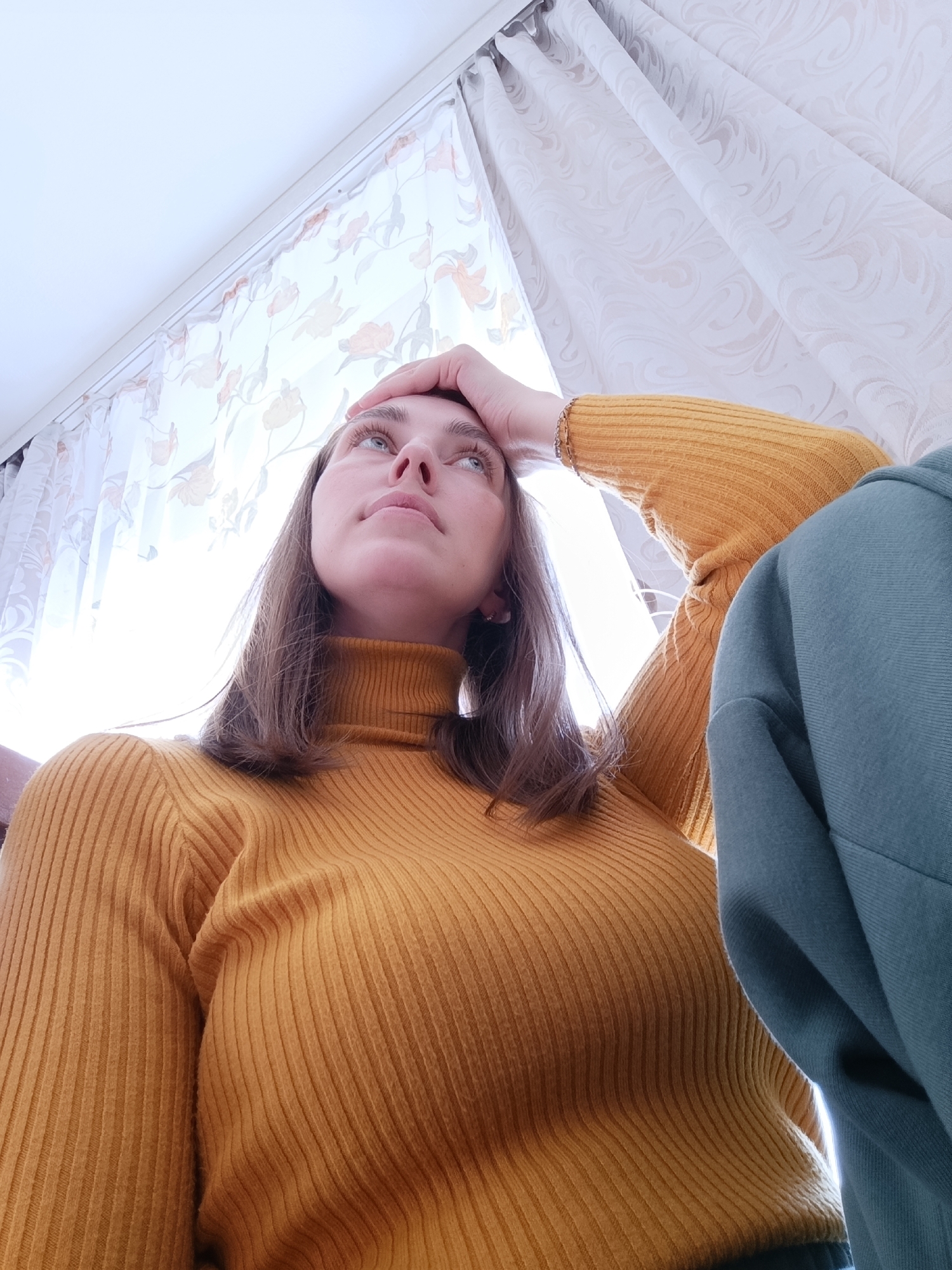 A low-angle shot depicts a fair-skinned young woman with shoulder-length brown hair wearing a mustard-yellow turtleneck sweater. Her left hand is raised, with fingers resting on her forehead. She is looking upwards with a contemplative expression. A portion of a blue-green garment or fabric is visible on the right side of the frame. The background features a white curtain with a light-colored floral pattern. Based on the available information, the location cannot be determined to be Фоки, Russia.