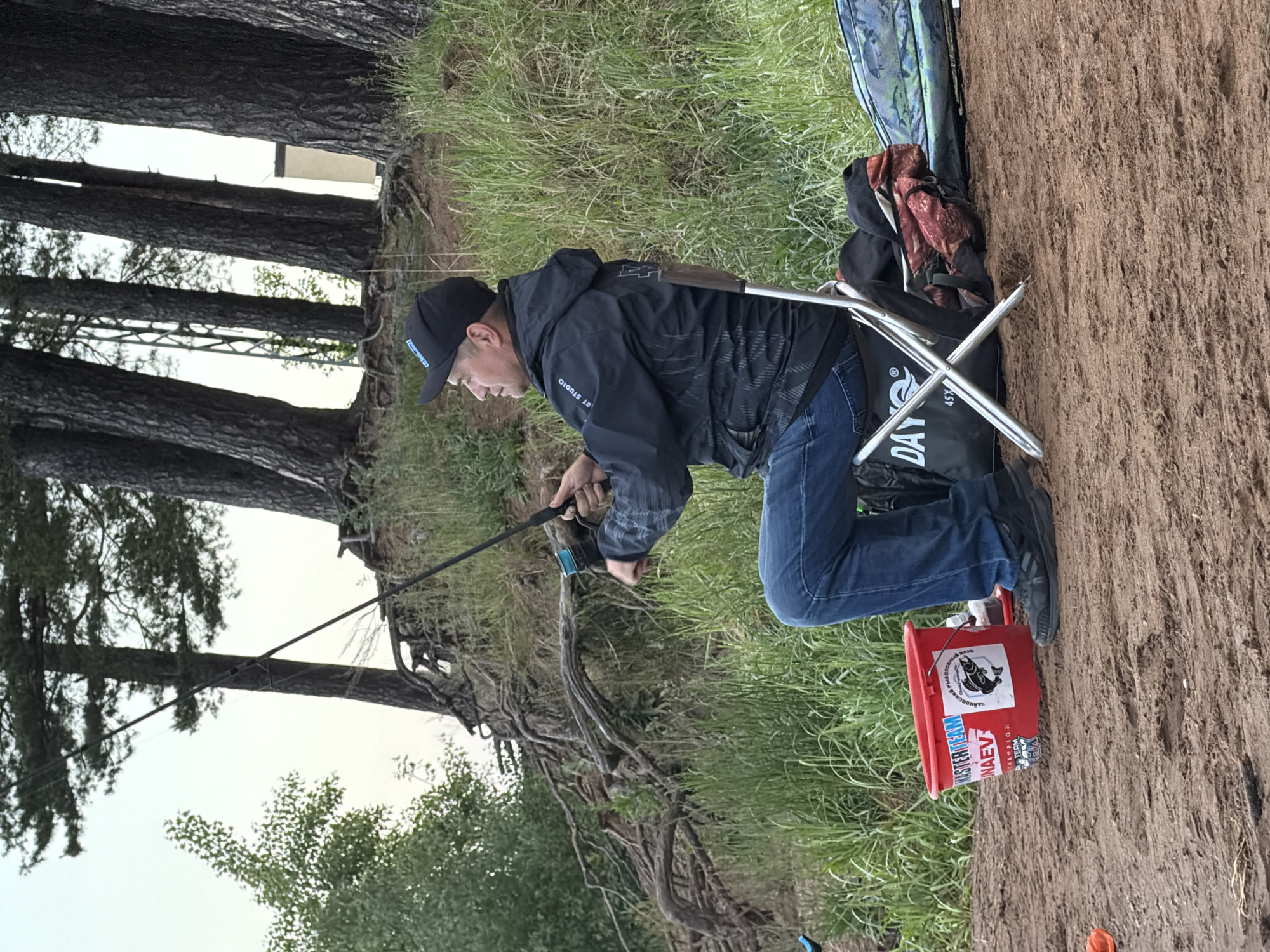 The image captures an outdoor fishing scene, likely on a riverbank or lake shore in Chaikovsky, Russia. An adult male is prominently featured, kneeling or sitting on a portable chair, actively holding a fishing rod and looking intently towards an unseen body of water. He is dressed in a dark baseball cap with an indistinct logo, a dark waterproof jacket that displays "STUDIO" on the sleeve and possibly "DAY" on the chest, blue jeans, and dark sneakers.
Behind the individual, a silver foldable camping chair is set up, draped with a dark bag that reads "DAY" and "45X". A backpack with a red and patterned design rests on the chair. To the man's right, a red plastic bucket sits on the ground, adorned with a black fish logo and various labels. Visible Cyrillic text includes "MASTERN TEAM," "МАЕВ" (Maev), and "РЫБОЛОВНЫЙ КЛУБ" (Fishing Club).
The immediate ground is sandy and earthy, gradually giving way to a dense patch of green grass further back. A line of tall, dark-barked trees, possibly pines or firs, stands on an eroded bank in the background, with their robust roots visible. Beyond the trees, an overcast, light sky is seen, suggesting either an early morning or late afternoon setting. The overall atmosphere appears calm and focused on the fishing activity.