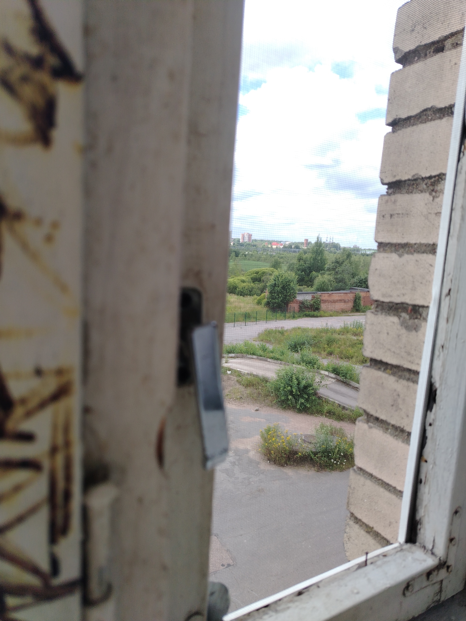 The image shows a view through an open, weathered window frame with a white painted exterior. The interior side of the window frame, on the left, is a light beige color with some dark, abstract markings. A metal window latch is visible on this side. The exterior view shows a cloudy sky with patches of blue. To the right, a portion of a brick wall is visible, constructed of light-colored bricks with visible mortar. The brick wall appears to be part of the building the window is in.
Beyond the window, a landscape unfolds. A gray asphalt road curves through a grassy area dotted with trees and bushes. In the middle ground, there is a low-lying brick building with a red roof, partially overgrown with vegetation. Further in the distance, a few more buildings, including what appears to be a tall, cylindrical structure and several residential buildings with red roofs, are visible on the horizon. The overall impression is a somewhat industrial or semi-rural setting. There are no people or animals visible in the image. The scene is likely in a non-urbanized area of Vyborg, Russia, possibly a residential area or an industrial periphery given the presence of the brick building and distant industrial-looking structures.