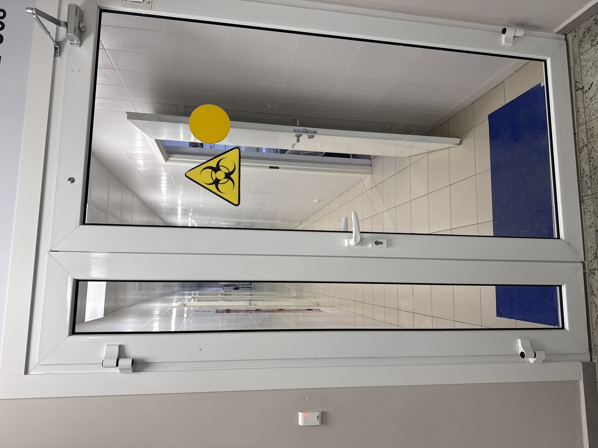 A white-framed window with a biohazard symbol is present. The window is likely located in a laboratory or medical facility. The reflection shows an interior with tiled walls and ceiling, indicating a sterile environment. A blue object is also visible through the window. The presence of a biohazard symbol indicates the potential for hazardous materials or biological agents within the reflected space. The city of Moscow, Russia is not visibly indicated within the image, hence the location cannot be confirmed.