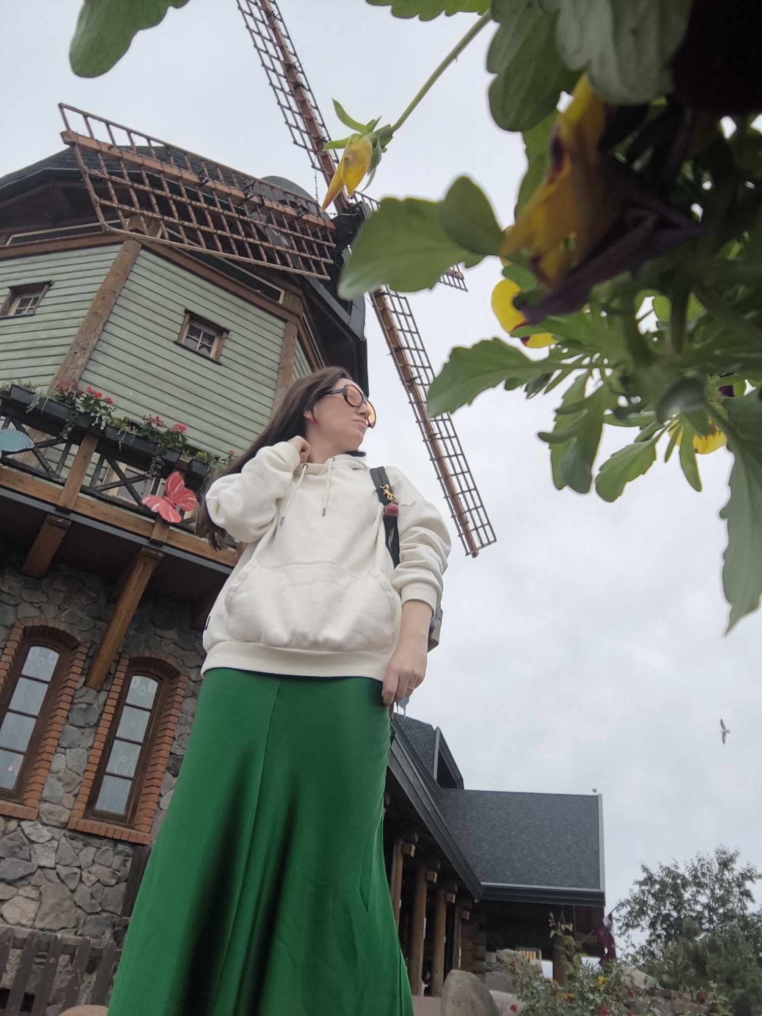A low-angle, medium shot captures a woman standing in front of a quaint, multi-story building designed to resemble a windmill, possibly a themed attraction in Riga, Latvia. The woman, seen from the waist down and partially from the side, wears a white hoodie and a long, flowing green skirt. She has her right hand raised to her neck and is looking off to her right. Behind her, the windmill-style building features a weathered wooden exterior with visible planks, small square windows, and decorative flower boxes along a balcony. The windmill's blades are visible at the top of the structure. In the foreground, blurred green leaves and yellow and purple flowers obscure the bottom right portion of the frame. The sky is overcast.