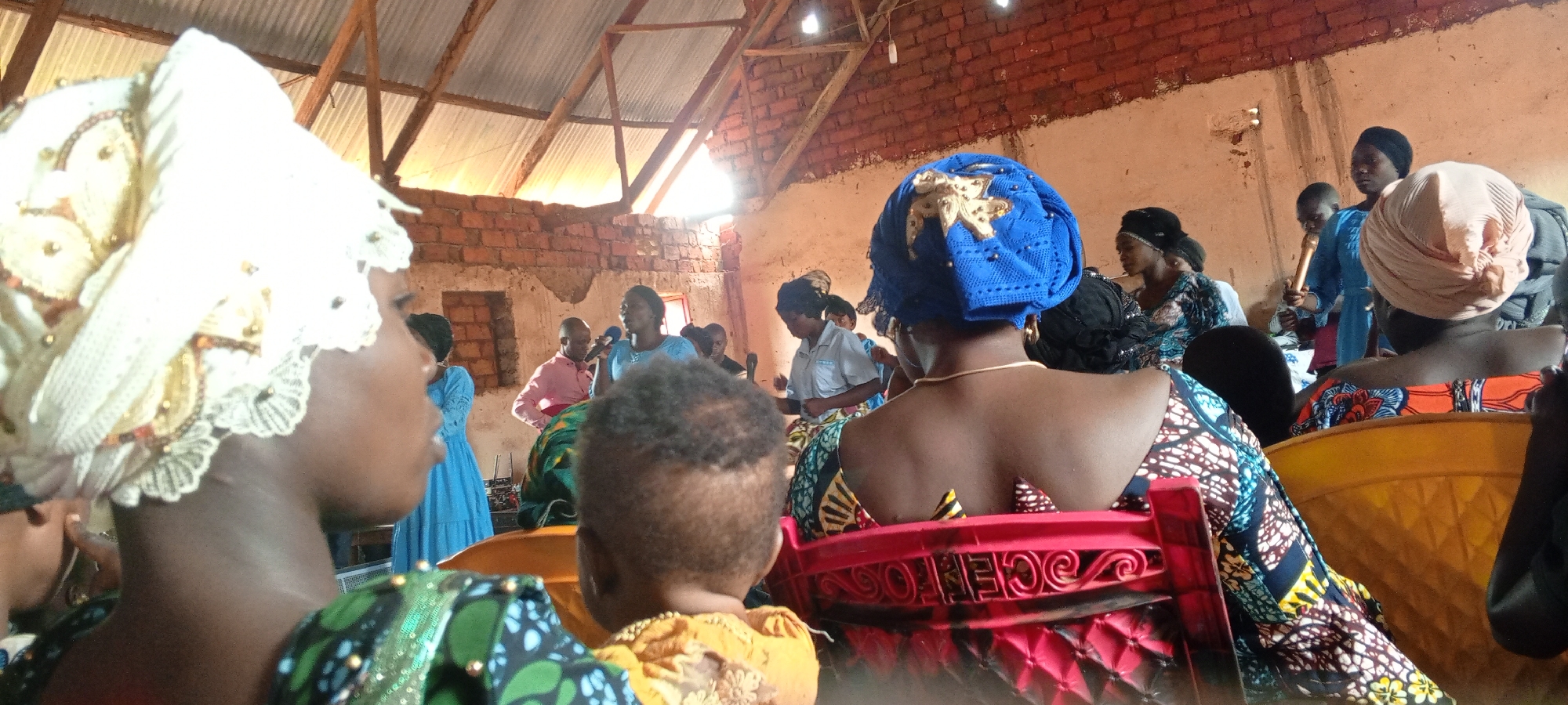 In Ngalula, Democratic Republic of the Congo, the photograph depicts a gathering of people inside a simple structure, likely a community hall or a church. The building appears rudimentary, with exposed brickwork and an open roof structure supported by wooden beams. Natural light streams through gaps in the roof.
Attendees are predominantly women, many wearing colorful, patterned clothing and distinctive head wraps. A child in yellow sits in the foreground, facing the same direction as the seated women. Several people stand and are interacting, some holding microphones. A man in a pink shirt stands near someone speaking. Others are dressed in lighter blue clothing. The focus is on the attendees and the sense of community within this unadorned setting.
