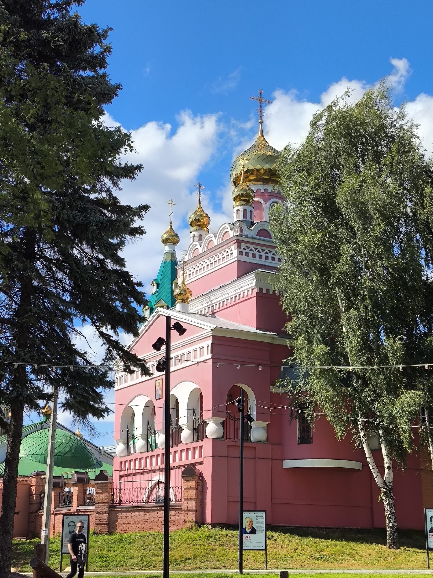 The image shows a bright pink orthodox church with golden domes. The church has multiple arched windows and decorative white trim. In the foreground, there are trees on both sides of the frame, with lush green leaves. The sky is bright blue with scattered white clouds. A person wearing a black t-shirt and dark pants is walking past a brick fence in front of the church. There are also posters displayed on stands near the fence. The overall scene suggests a sunny day in a city. The church is the Alexander Nevsky Cathedral, located in Izhevsk, Russia.
