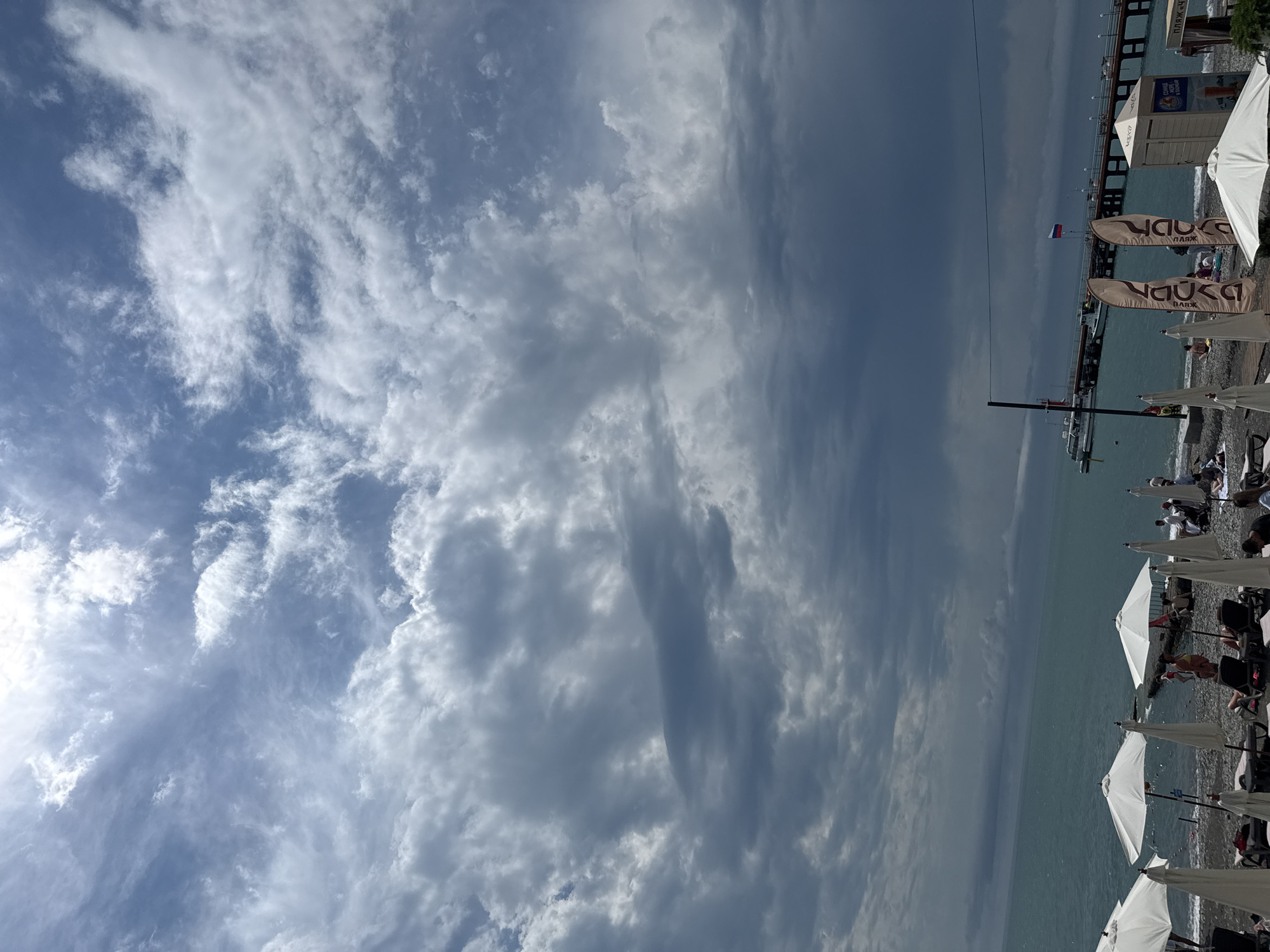 The image depicts a cloudy day at a beach in Sochi, Russia. In the foreground, several white beach umbrellas are set up, with lounge chairs placed underneath them. Some people are visible on the beach, relaxing or walking. The sea is a turquoise color, and a long pier extends into the water. A Russian flag is flying from the pier, and a boat is docked there. In the background, the sky is filled with dramatic clouds, suggesting a possibility of rain. The overall scene conveys a typical beach day in a coastal city.
