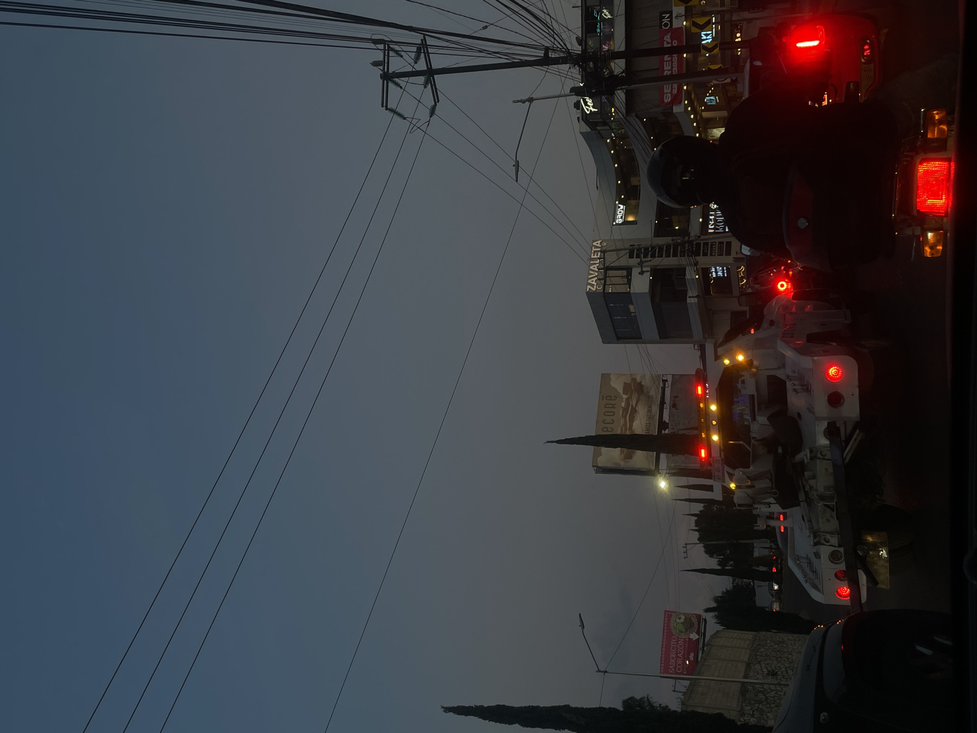 Here's a description of the image:
The photograph captures a street scene in Puebla, Mexico, at dusk or night. A prominent sign reads "Zavaleta". Utility poles and numerous overhead wires dominate the upper portion of the frame, crisscrossing against a dark sky. In the foreground, a motorcycle with a rider is visible, brake lights illuminated, and traffic is congested. Further down the street, there is a large, white commercial truck with its taillights illuminated, followed by other vehicles. Billboards advertising local businesses are visible on the sides of the road. The scene has a busy, urban feel.