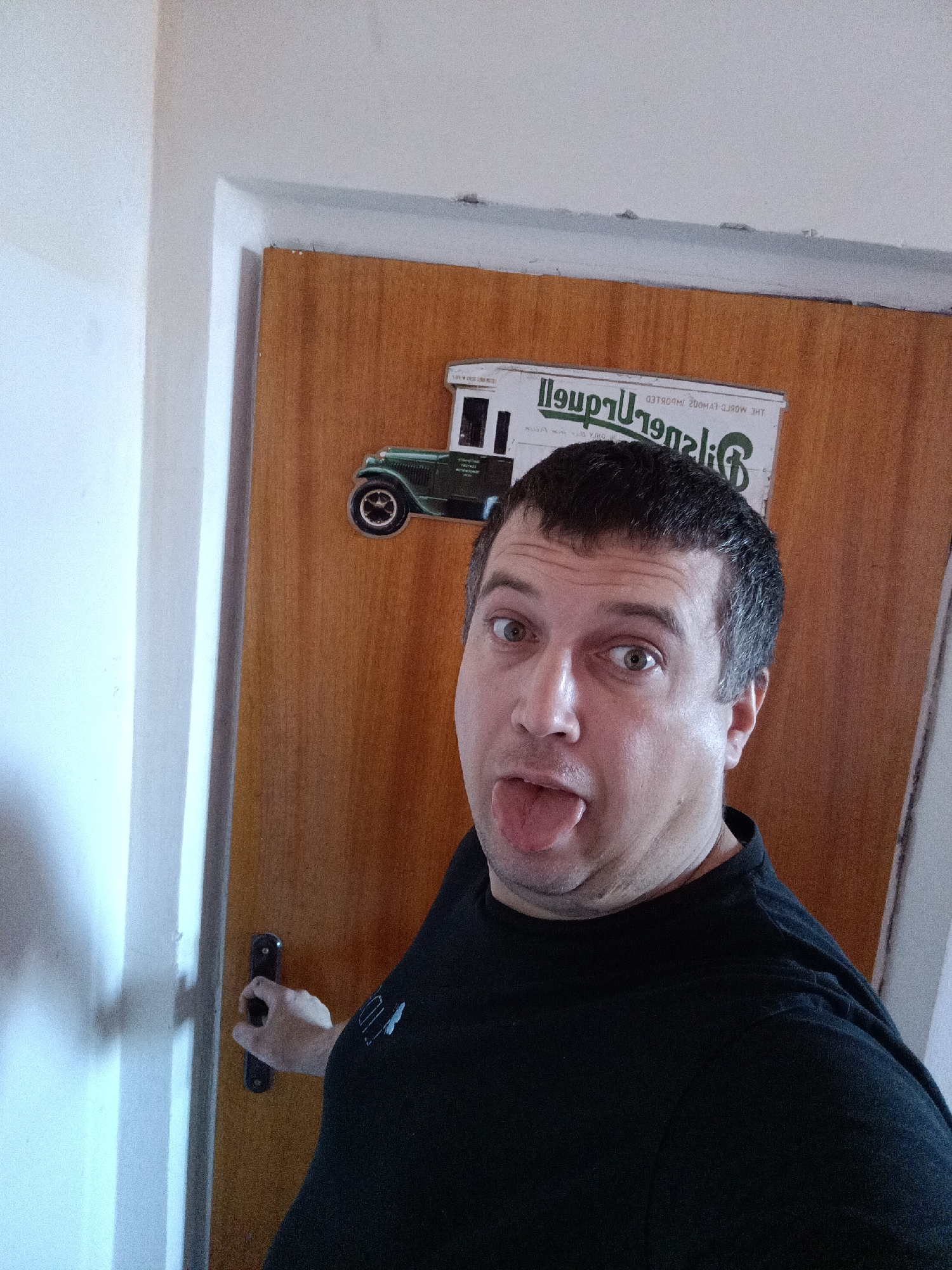 An adult male subject is depicted in a close-up, waist-up, selfie-style photograph, taken indoors in the city of Dalovy, Czechia. The subject, who has short, dark hair and light eyes, is wearing a black crew-neck t-shirt. He is positioned in front of a wooden door, which features a dark rectangular handle held by his right hand. His head is slightly tilted, and he is looking directly at the camera with wide eyes and his tongue extended in a playful or silly expression.
Behind the subject, a light-brown wooden door is visible, bordered by white trim. Affixed to the upper portion of the door is a rectangular sign or decal. The sign features a graphic of a vintage green delivery truck facing left, and mirrored text. When corrected for mirroring, the text appears to read "THE WORLD-FAMOUS" in smaller white letters, and "Gilgenberg" in larger green letters, with additional obscured text below. To the left of the door, a plain white wall is visible. The immediate environment appears to be an interior space, likely a hallway or room, characterized by simple architectural elements.