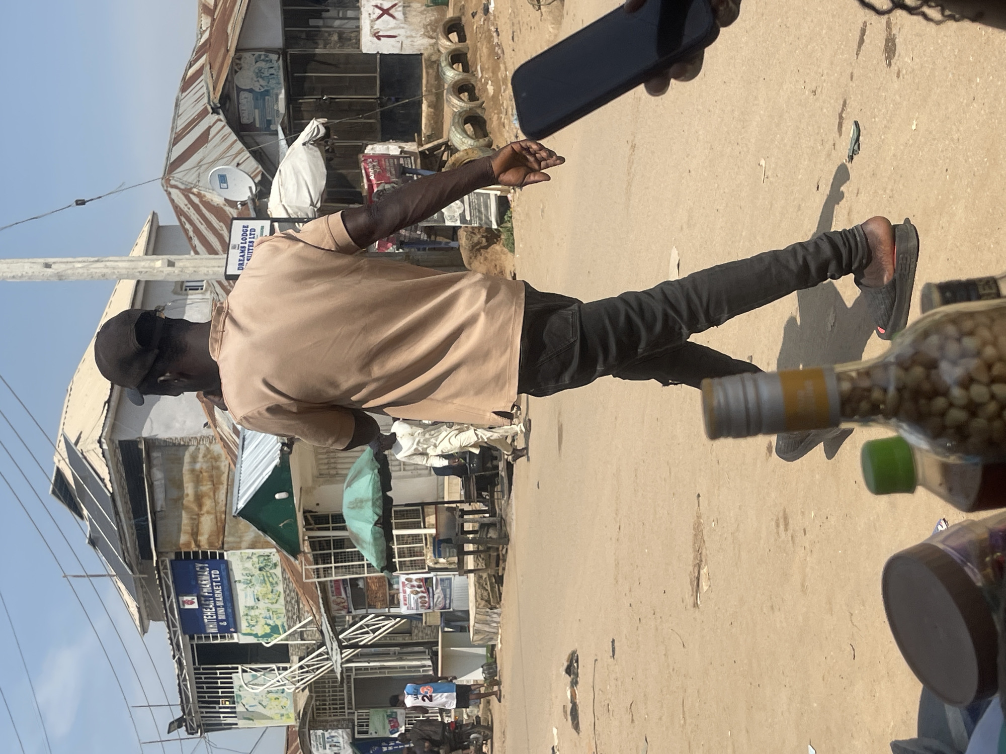 A man wearing a tan t-shirt and black pants walks across a dusty, unpaved street in an outdoor market. He has a black baseball cap on his head and is looking down and to his right. His left arm is extended. Behind him, a building with a sign that reads "WHITEHEART PHARMACY & MINI-MARKET LTD" is visible. Other structures, including a building with "DREAMS LODGE SUITES LTD" on a sign, are also present. Several people are in the background, some walking and others near stalls. There are umbrellas, tables, tires, and various objects scattered on the ground. In the foreground, a bottle filled with nuts and a jar are visible. The sky is clear and blue. The scene appears to be in Abuja, Nigeria.