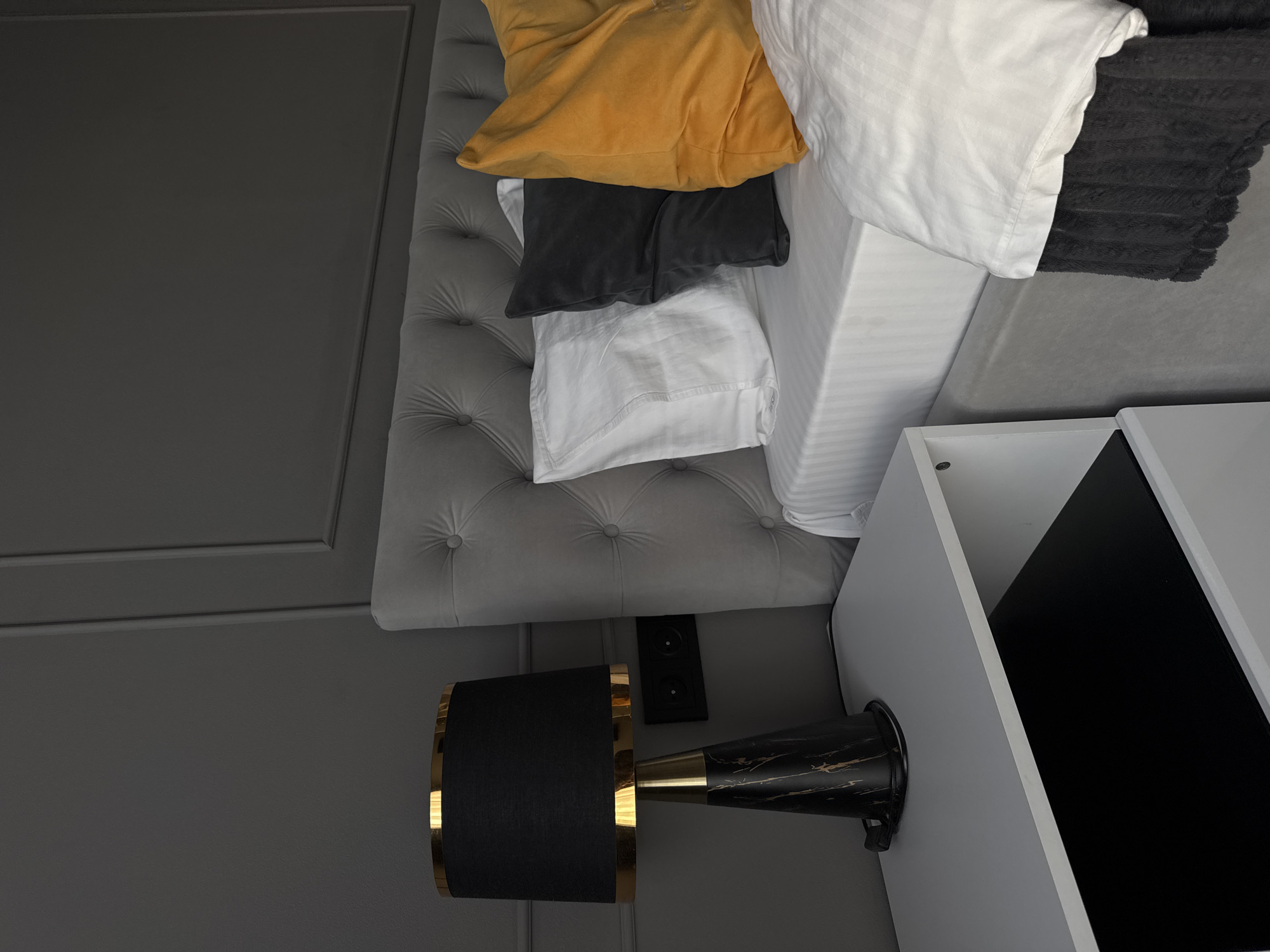 The image shows a section of a bedroom interior. The room features a gray, tufted headboard and gray walls with decorative molding. A bed is made with white striped bedding, a white pillow, a yellow decorative pillow, and a gray decorative pillow. A black textured throw blanket rests at the head of the bed. Adjacent to the bed is a white nightstand with a black inset. A black and gold lamp is positioned on the wall next to the headboard. A double electrical outlet is mounted to the wall.
There is no information available to indicate the scene takes place in Volgograd, Russia.