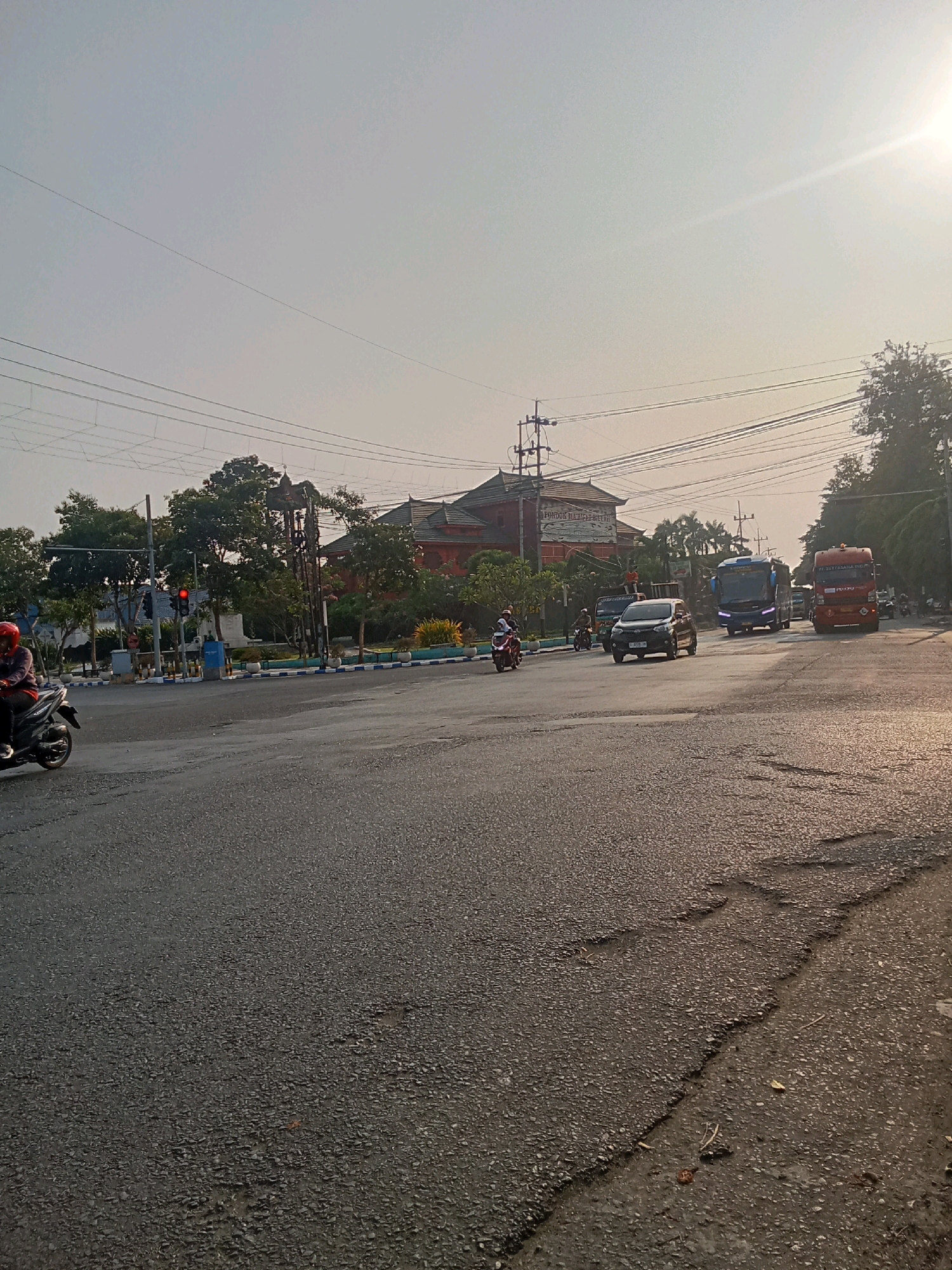 The image captures a street scene in Kaliwungu, Indonesia, bathed in the warm light of a setting or rising sun, casting long shadows and creating a bright glare in the upper right corner. The road surface is asphalt, showing signs of wear with patches and cracks. In the foreground, the road stretches towards the horizon.
On the left side of the road, a motorcycle rider wearing a red helmet is visible, traveling on the asphalt. Further down the road, a traffic light displays a red signal. Adjacent to the traffic light is a pole with overhead electrical wires. Behind the traffic light, a building with a traditional Indonesian architectural style, characterized by its dark tiled roof, is partially visible. Green trees line the side of the road, with some foliage overhanging the street.
In the middle ground, several vehicles are present. A dark-colored sedan is in motion. A blue bus with a white stripe and a red truck are also on the road. Several other motorcycles are scattered throughout the scene. The background consists of more trees, buildings, and utility poles, all silhouetted against the hazy sky. The overall impression is of a moderately busy intersection in a tropical urban environment.