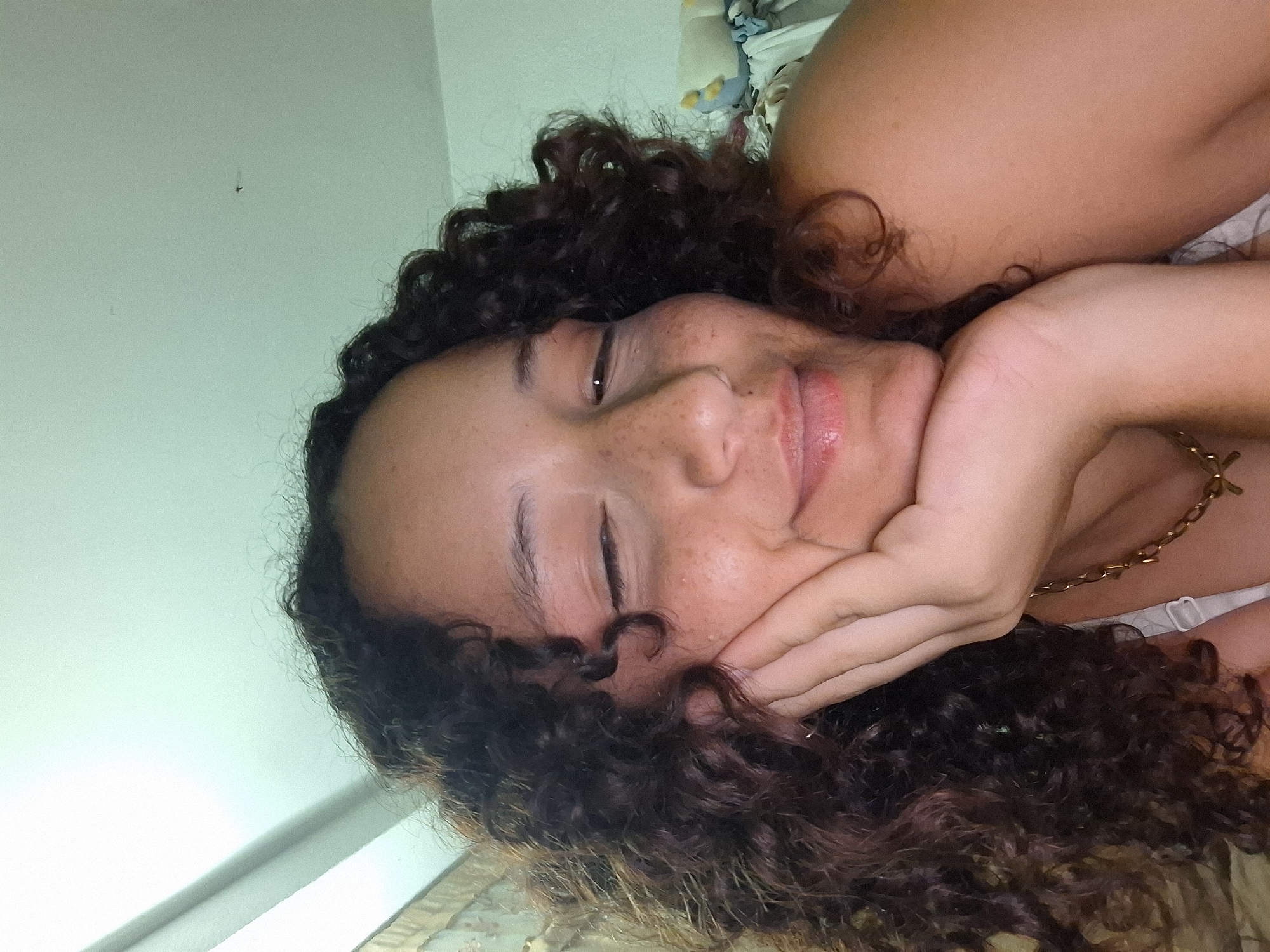 A close-up selfie shows a young woman with curly dark brown hair, leaning her head on her hand. Her eyes are closed, and she has a slight smile. She has light skin with freckles across her nose and cheeks. She is wearing a thin gold chain necklace. The background is blurred, but appears to be an indoor setting with a light-colored ceiling. The image quality is slightly grainy, suggesting it was taken with a mobile phone. The context of Juan Griego, Venezuela is not discernible from the visual information provided in the image.