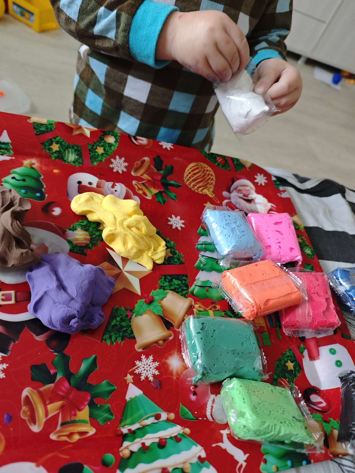 The image captures a close-up, high-angle view of a child playing with colorful modeling clay or play-doh, set in what appears to be a home in Markovskii, Russia. The child, wearing a brown and light blue checkered long-sleeved top with blue cuffs, is visible from the chest down, with their hands centered in the upper portion of the frame. They are holding a small, clear plastic package containing white modeling clay, possibly in the act of unwrapping it.
The play takes place on a vibrant red mat or tablecloth adorned with festive Christmas illustrations, including Santa Claus, snowmen, various Christmas trees, golden bells, snowflakes, and wreaths. Several pieces of modeling clay are scattered across this festive surface. Three colors are unwrapped and in various forms: a large blob of bright yellow clay, a smaller pile of brown clay, and a mass of purple clay. Additionally, numerous other colors remain neatly packaged in clear plastic, including bright blue, hot pink, orange, dark green, light green, and more hot pink and blue blocks. These packaged blocks are primarily grouped on the right side of the mat.
The scene suggests an engaging and creative playtime activity. The lighting is consistent with an indoor setting, likely daytime, though no specific time cues or windows are visible. The focus is entirely on the child's hands and the materials, conveying a sense of concentration and playfulness. There is no visible text in the image.