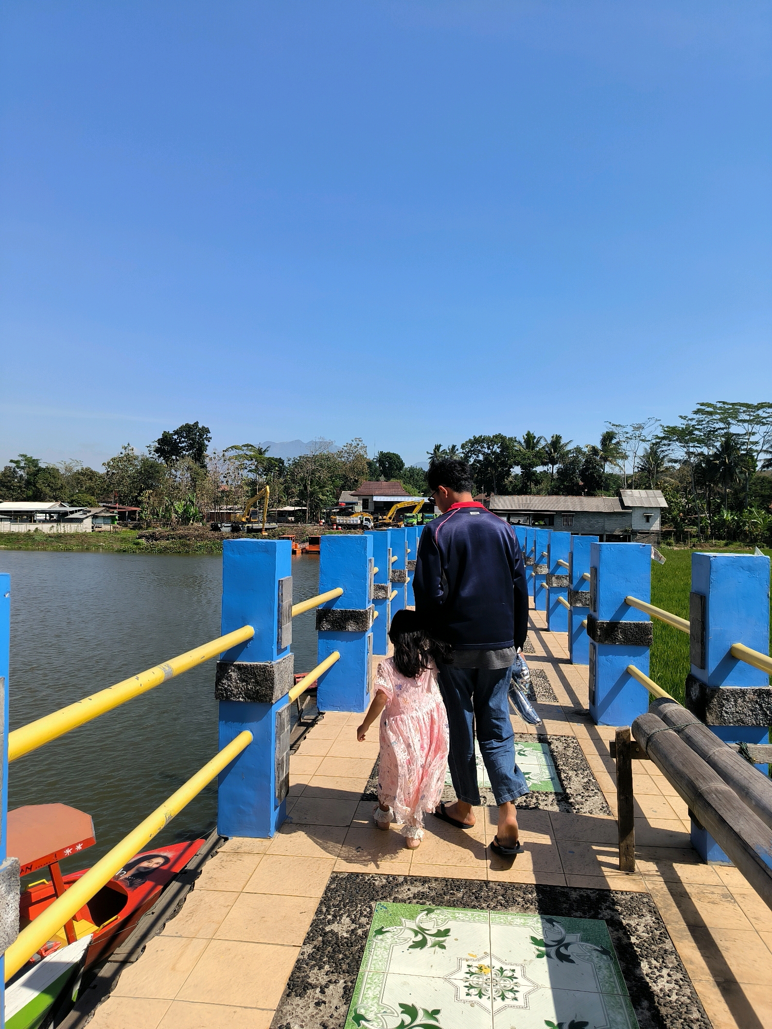 The image depicts a father and daughter walking on a pathway adjacent to a body of water under a clear blue sky. The pathway is bordered by a blue railing with yellow poles. In the background, there are trees, buildings, and some construction equipment. The setting appears to be outdoors, possibly a park or a waterfront area. The location is identified as the city of Tuntang, Indonesia. The father is wearing a dark blue jacket and jeans, while the daughter is in a pink dress. They are walking away from the camera.