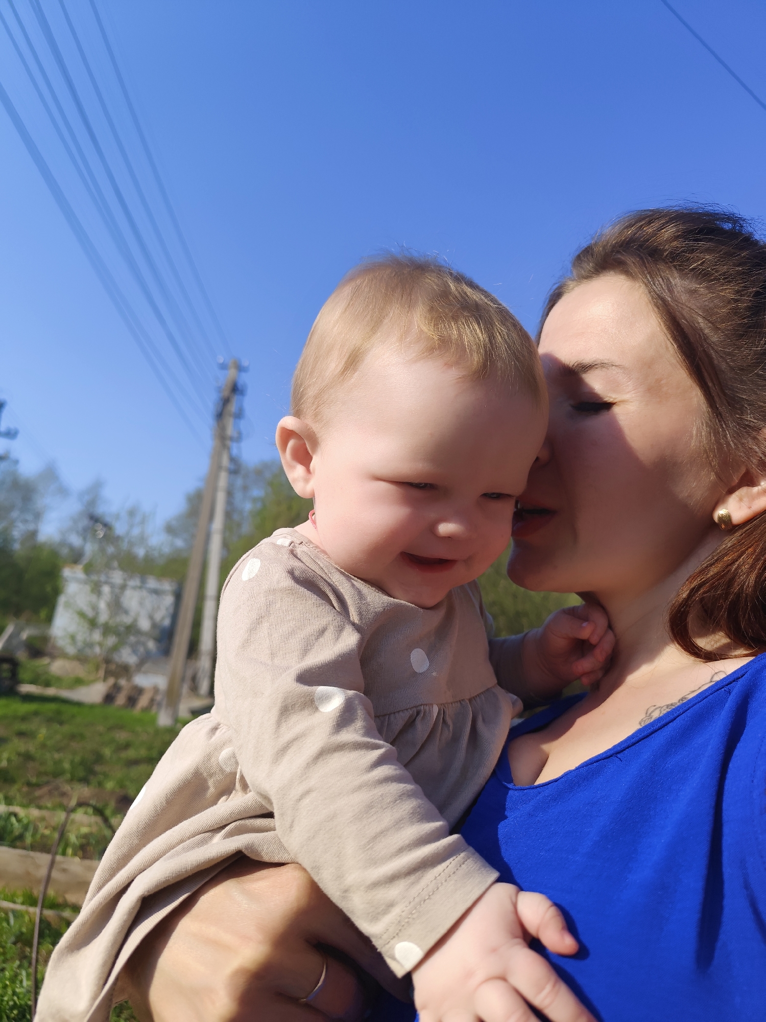 Here's a detailed description of the image:
**People:** A woman is holding a baby. The baby is fair-skinned with blonde hair, wearing a light brown, long-sleeved dress with white polka dots. The baby is smiling slightly and looking down. The woman has brown hair pulled back and is wearing a blue top. She has her eyes closed and appears to be nuzzling the baby.
**Environment:** The setting is outdoors on a sunny day, suggested by the clear blue sky and the shadows. The background includes greenery, possibly a garden or park. In the far distance, there is a utility pole with power lines, and the corner of a light-colored building.
**Action/Interaction:** The central interaction is the woman holding and seemingly kissing the baby.
**Location Clues:** There is not any readily apparent visible information from the image that leads to the city of Фоки, Russia.