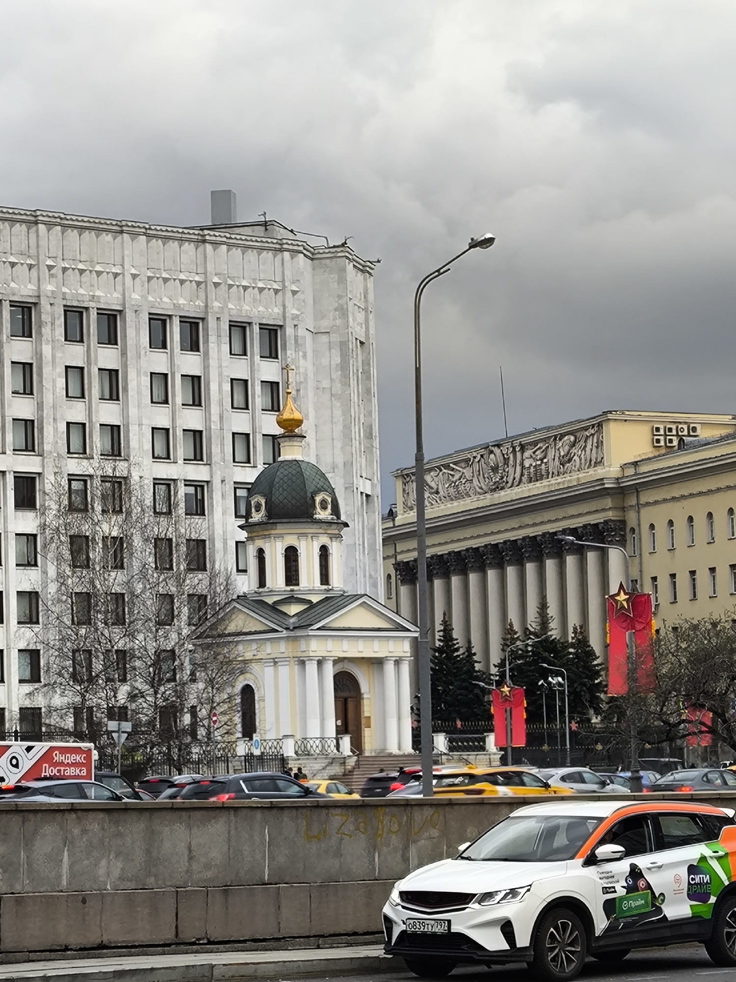 An urban scene in Moscow, Russia, is depicted under an overcast, grey sky. The view is dominated by several distinct buildings and a busy street with traffic.
In the mid-ground, a small, classical-style chapel-like building with a green dome topped by a golden cupola and cross stands prominently. Behind it, to the left, is a tall, light-colored, multi-story building with numerous windows, exhibiting a stern, blocky architectural style. To the right of the chapel, a grand, yellow building with a colonnade and an elaborate sculptural frieze along its roofline is visible. Red banners with gold stars hang in front of this building, suggesting a festive or commemorative occasion.
The foreground features a concrete barrier separating the viewer from a multi-lane road. Several cars are visible, indicating traffic congestion. A white SUV, possibly a taxi, with orange and green livery is prominently parked or moving slowly on the right. Another vehicle, a white delivery van with "Яндекс Доставка" (Yandex Delivery) written in red, is partially visible behind the barrier on the left. A tall street lamp extends into the sky from the right side of the image.
Visible text includes "Яндекс Доставка" on the delivery van. On the white SUV, "СИТИ" (CITY) and "О пройти" are partially visible, along with the Russian license plate "0839ТУ797". The overall atmosphere is that of a bustling city on a cloudy day.