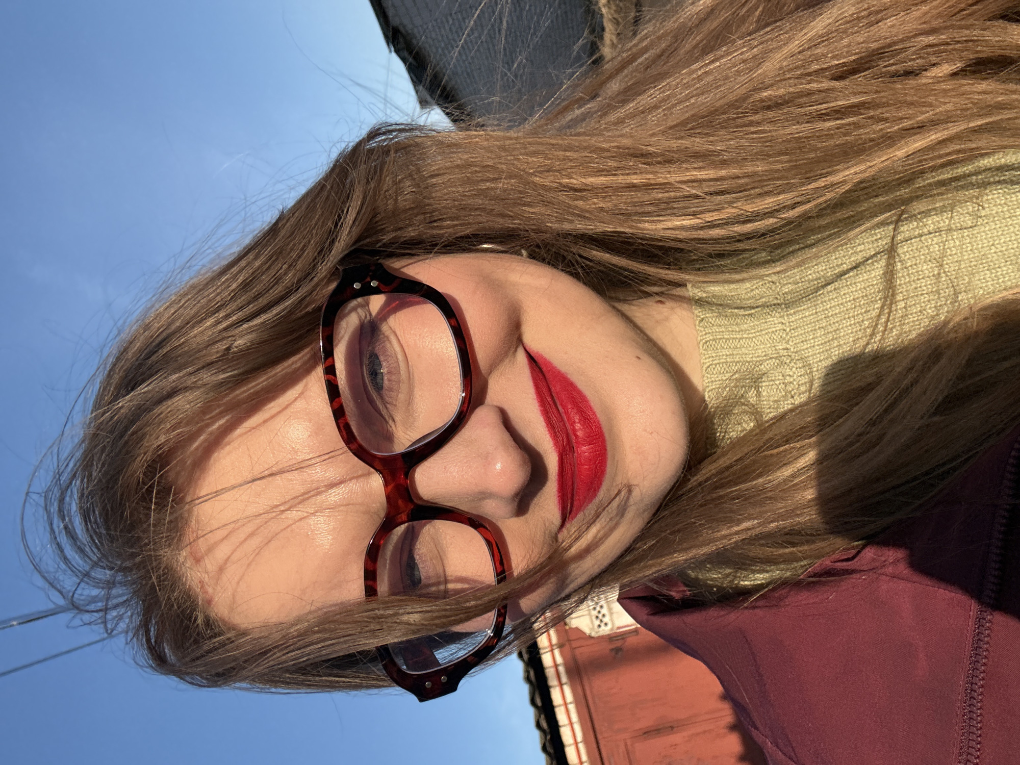 A woman with long brown hair and red lipstick is wearing red-framed glasses. She is wearing a light green sweater and a dark red jacket. The background includes a blue sky and parts of buildings, possibly in the city of Izhevsk, Russia, based on visual clues and contextual assumptions. The image appears to be taken from a low angle.