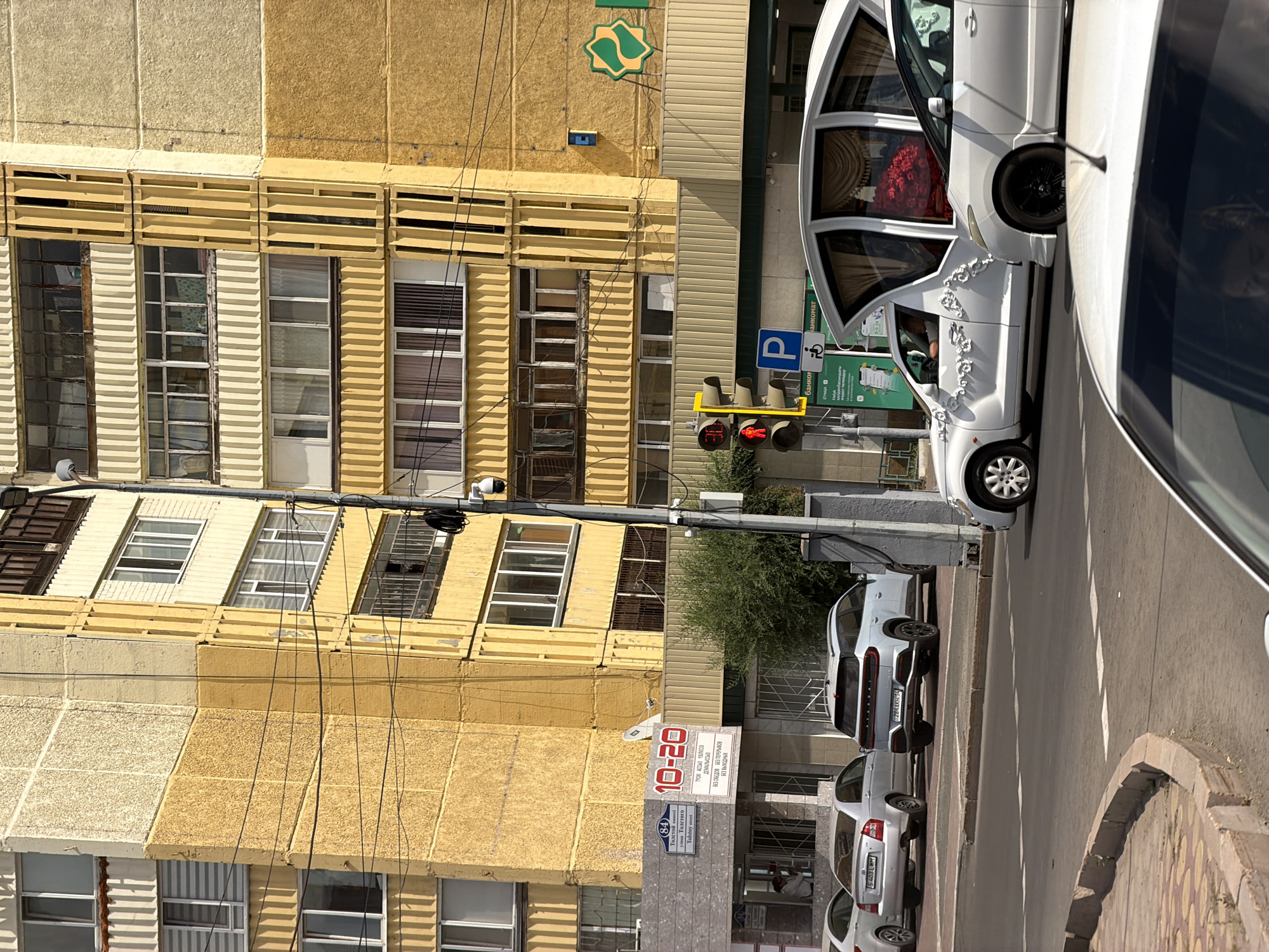The image depicts a street scene in Pavlodar, Kazakhstan, under a clear sky. A yellow apartment building dominates the background. In the foreground, cars are visible on the road, including a white hearse adorned with floral decorations. Traffic lights are present, with one showing a red light and a pedestrian signal indicating to stop. Several commercial establishments are situated at the base of the apartment building, featuring signage in Cyrillic and Roman alphabets. The street is paved, with a concrete curb and sidewalk. The overall impression is a typical urban environment.