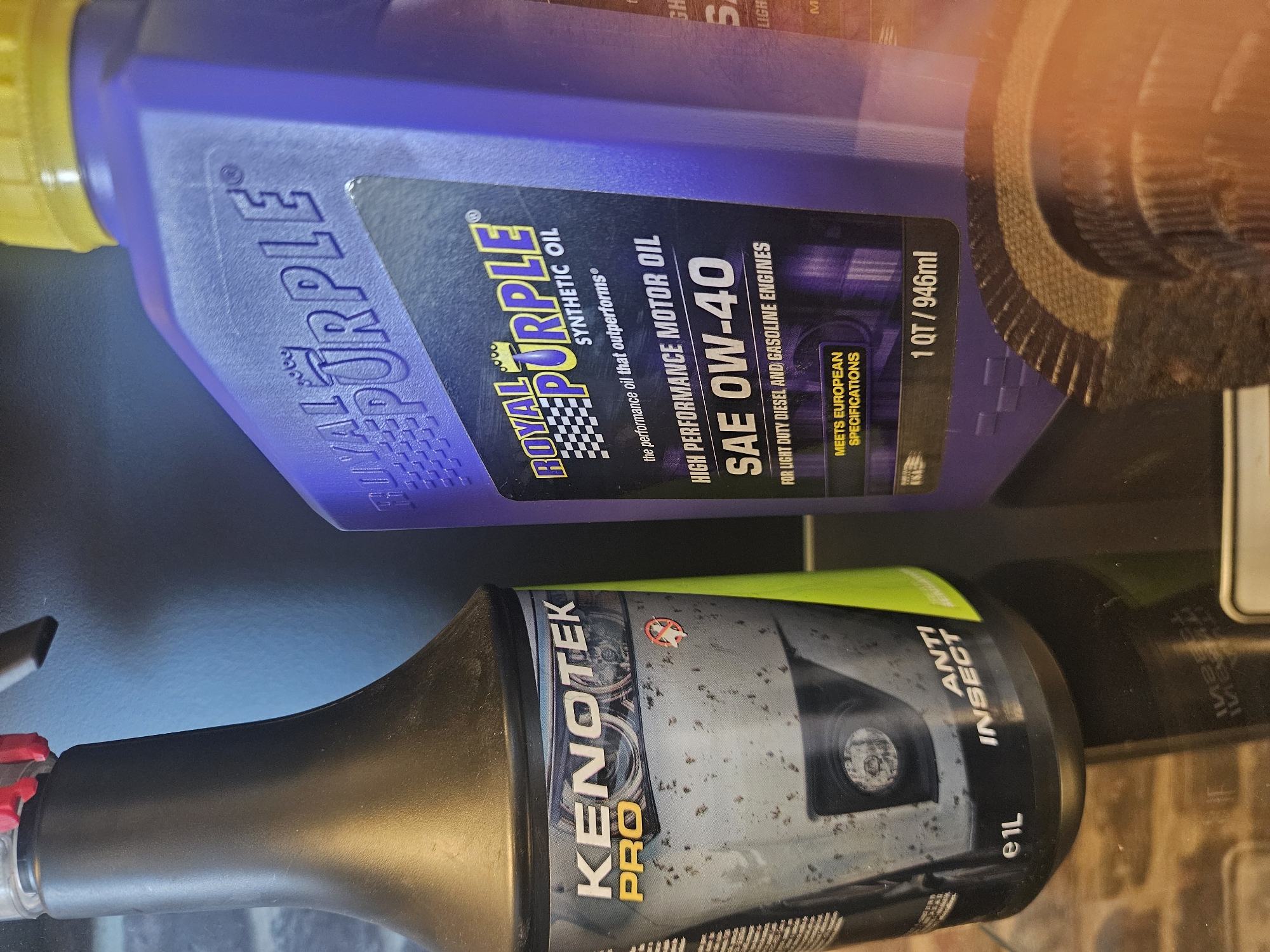 The image displays two bottles of automotive fluids placed on a surface, possibly a shelf or table. On the left is a black spray bottle with a lime green label featuring the brand "KENOTEK PRO" and text "ANTI INSECT" along with "e1L". The bottle has some smudges and marks on its surface. To the right is a purple bottle with a yellow cap, labeled "ROYAL PURPLE SYNTHETIC OIL". This bottle contains "HIGH PERFORMANCE MOTOR OIL SAE 0W-40 FOR LIGHT DUTY DIESEL AND GASOLINE ENGINES" and also states "MEETS EUROPEAN SPECIFICATIONS" and "1 QT / 946ml". A small portion of a cog or gear-like object is visible in the foreground, out of focus. The background consists of a brick-patterned wall and a darker, possibly blue, wall section. The scene appears to be an indoor setting. There are no people or animals visible in the image. No specific location in Bemmel, Netherlands can be identified from the image.