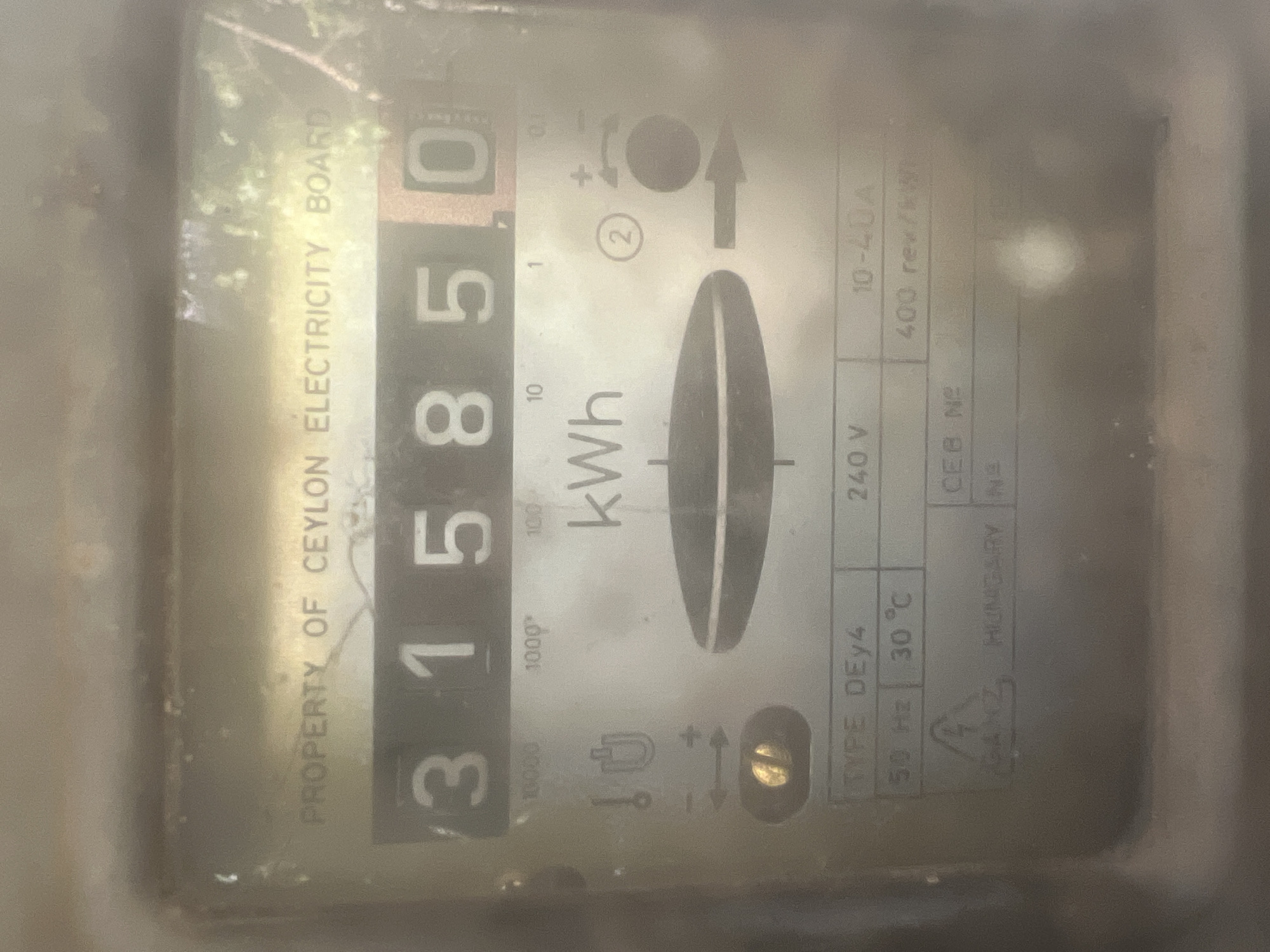 The image displays a close-up, slightly rotated view of an old, electromechanical electricity meter, likely enclosed within a protective cover that appears hazy, dusty, and scratched. No people are visible.
The primary subject is the meter's face, featuring a mechanical readout showing "315850". The final digit, "0", is on a distinct red background, suggesting it represents tenths of a kilowatt-hour. Below the main reading, smaller numerical indicators for 10000, 1000, 100, 10, 1, and 0.1 kWh are arranged vertically. To the right, a black and white spinning disk, characteristic of analog meters, is partially visible, indicating active electricity consumption. An arrow above the disk points to the right, showing the direction of rotation.
Numerous textual details and specifications are printed on the meter's face. Along the left edge, vertically aligned text reads "PROPERTY OF CEYLON ELECTRICITY BOARD," indicating the meter belongs to the electricity utility in Ceylon (now Sri Lanka). Other labels include "kWh" for the unit of energy, "TYPE DEy4", "240 V" (voltage), "50 Hz" (frequency), and "30°C" (operating temperature). The current range is specified as "10-40 A", and the calibration is "400 rev/kWh", indicating 400 revolutions of the disk per kilowatt-hour. The manufacturer "GANZ HUNGARY" is also visible, along with a partially visible "CEB Nº". A lightning bolt symbol is located near the manufacturer's name.
The meter's condition suggests significant age and prolonged use within its installed environment.