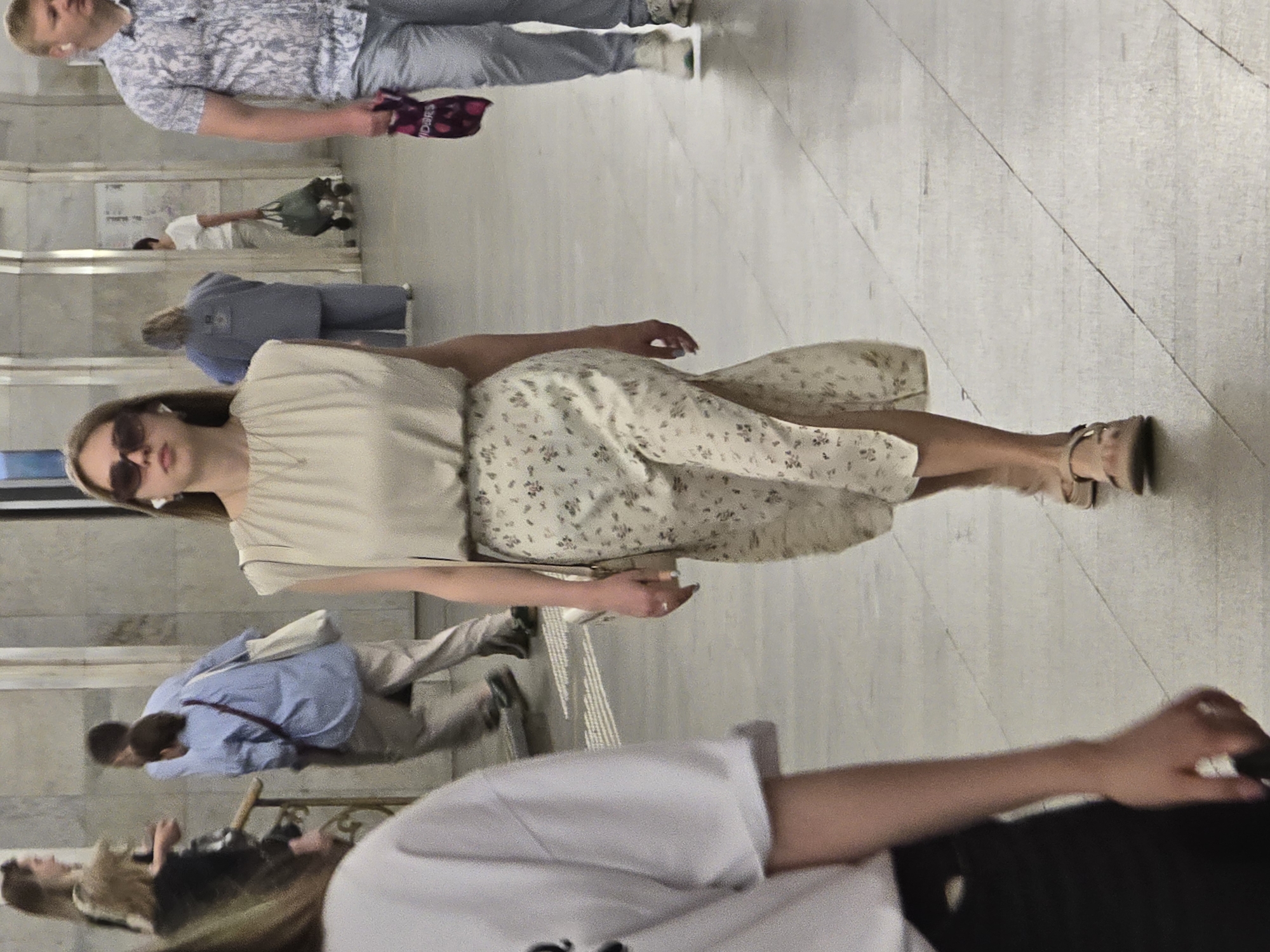 A full shot shows an indoor space in Unknown, Unknown. People are observed within this setting. A woman is standing, wearing sunglasses, a white top, and a floral print skirt. She is interacting with other individuals in the scene. Additional persons present include a man in a blue shirt and gray pants, another in a patterned shirt and gray pants, and others further in the background, some of whom are seated. The setting appears to be a public area, possibly a subway station or a similarly large public space given the architectural design. The flooring has a distinct pattern. The overall composition and placement of subjects indicate a candid shot of daily life activity.