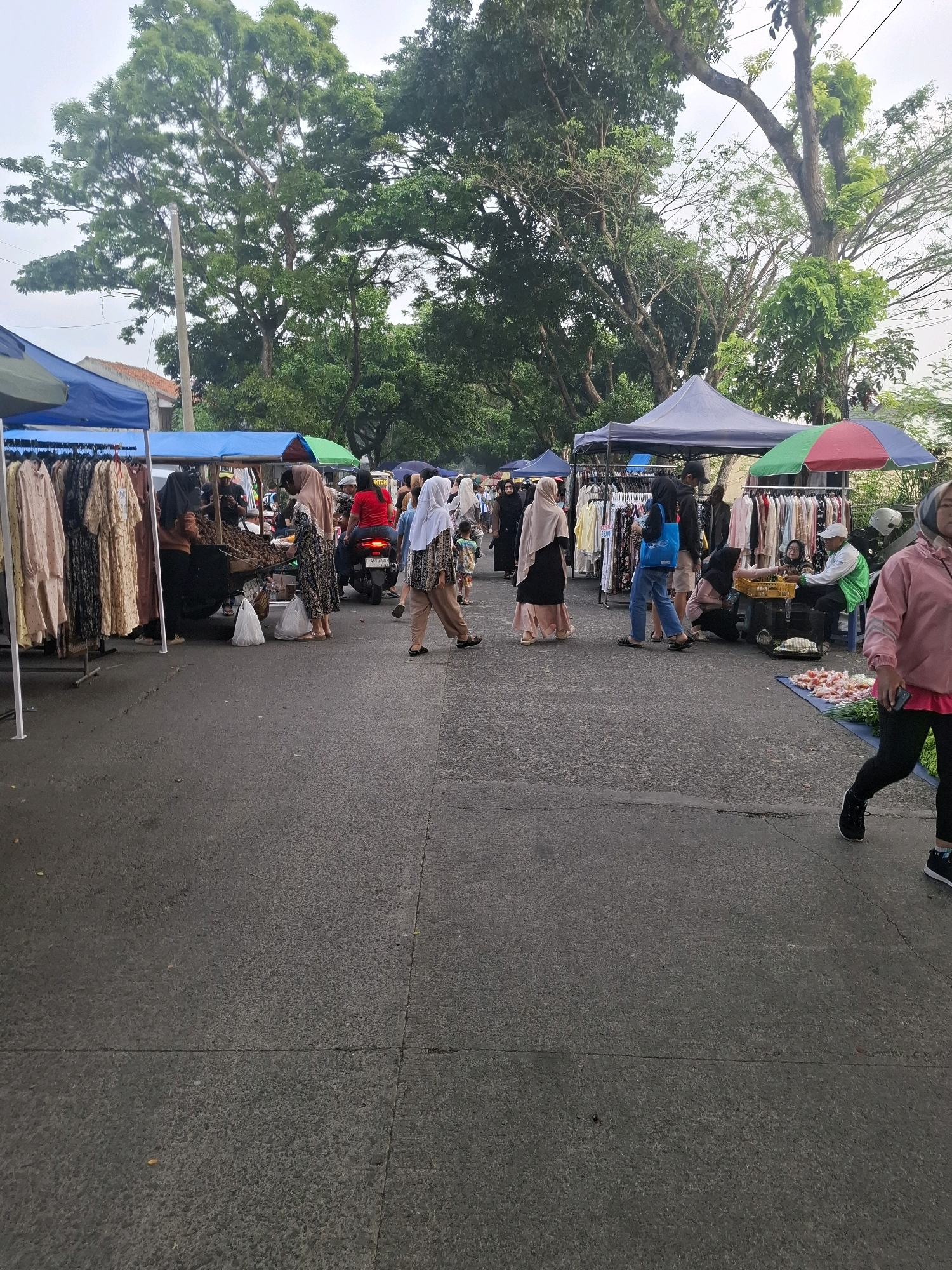 The image depicts a bustling outdoor market scene in Karangtengah, Indonesia. In the foreground, the asphalt surface of a road is visible, marked with a faint line indicating a lane or boundary. Several people are engaged in shopping or selling activities at various stalls. A woman in a pink jacket and black leggings is walking past, while another woman in black pants and a patterned top with a white headscarf is also walking.
To the left, a clothing stall is set up with racks of dresses on display. Several individuals are browsing or attending the stall. Further down the road, more stalls are visible, some with awnings providing shade. One stall appears to be selling produce, with a person attending it and a motorcycle parked nearby. Another stall has clothing racks and an umbrella providing shade.
The environment is characterized by mature trees lining the street, suggesting an urban or semi-urban setting with greenery. The sky is overcast, indicating potentially cloudy weather. The overall atmosphere is one of a lively marketplace with people actively participating in commerce.