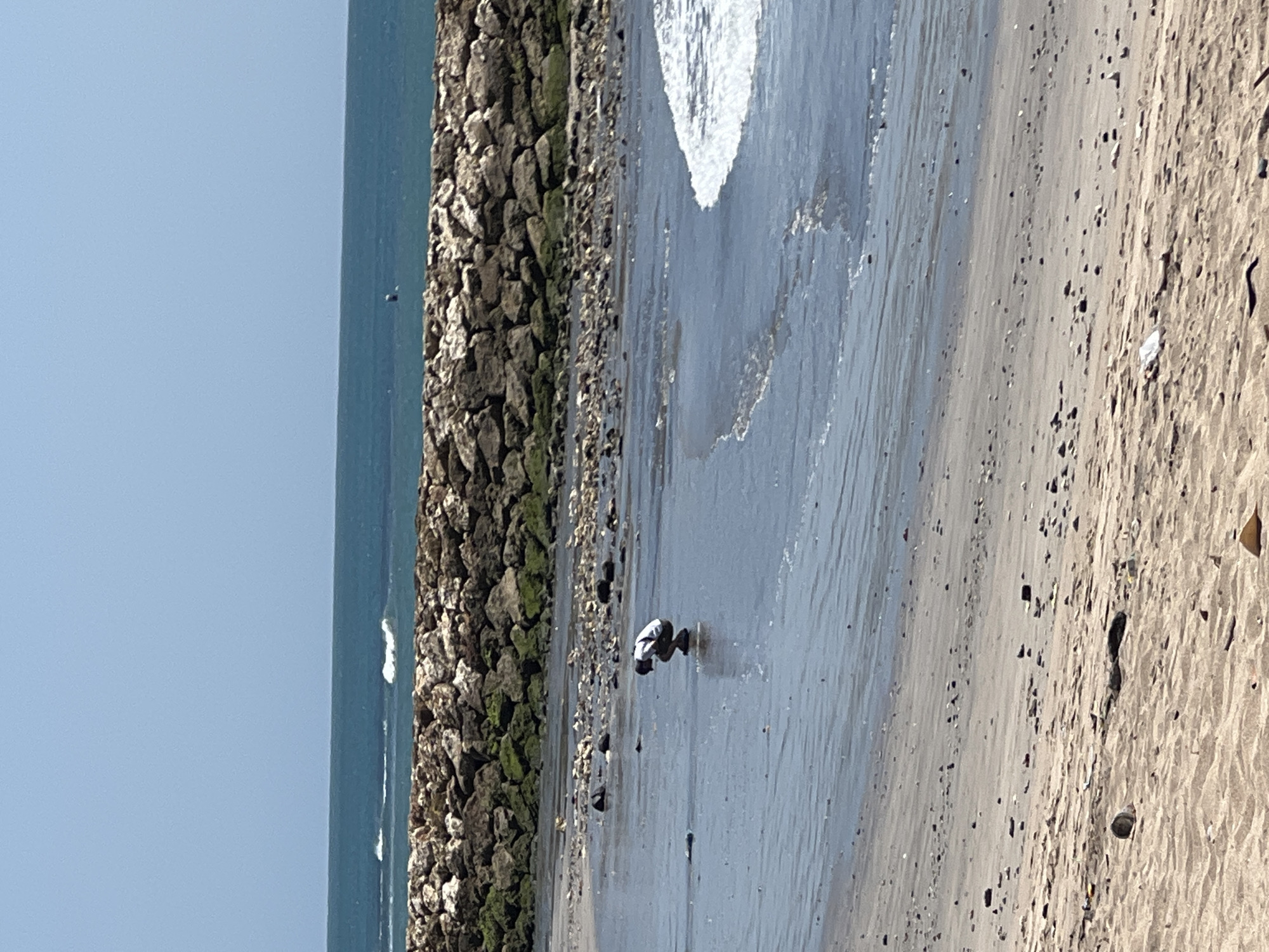 An individual is crouching on a wet sandy beach near the ocean. The person is wearing a white shirt and dark pants. The ocean is calm with small waves. In the background, a large rock wall extends into the ocean. The sky is clear and blue. The beach is scattered with small rocks and some debris. This scene is located in Kuta, Indonesia.
